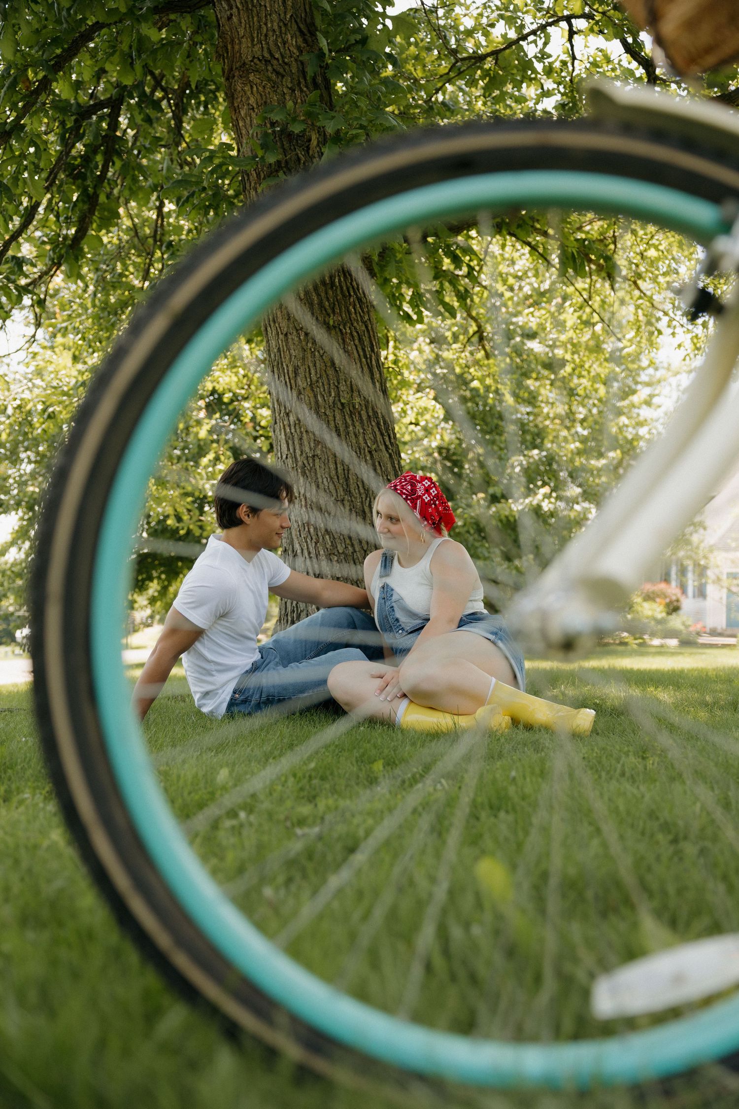 Couple sitting on grass under a tree viewed through a teal bicycle wheel in a sunny park.