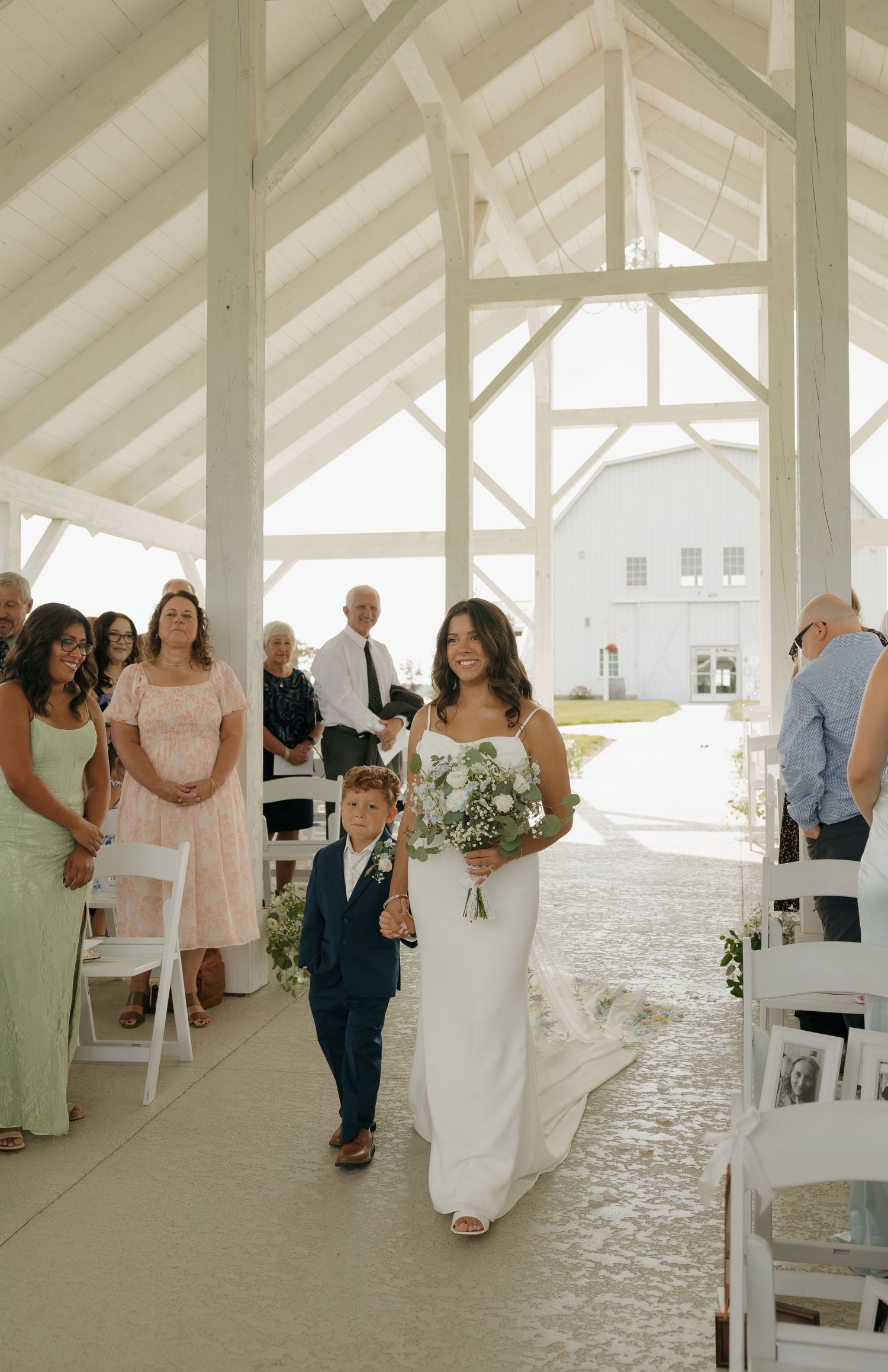 Bride walks down the aisle with a young ring bearer in a white open-air wedding chapel.