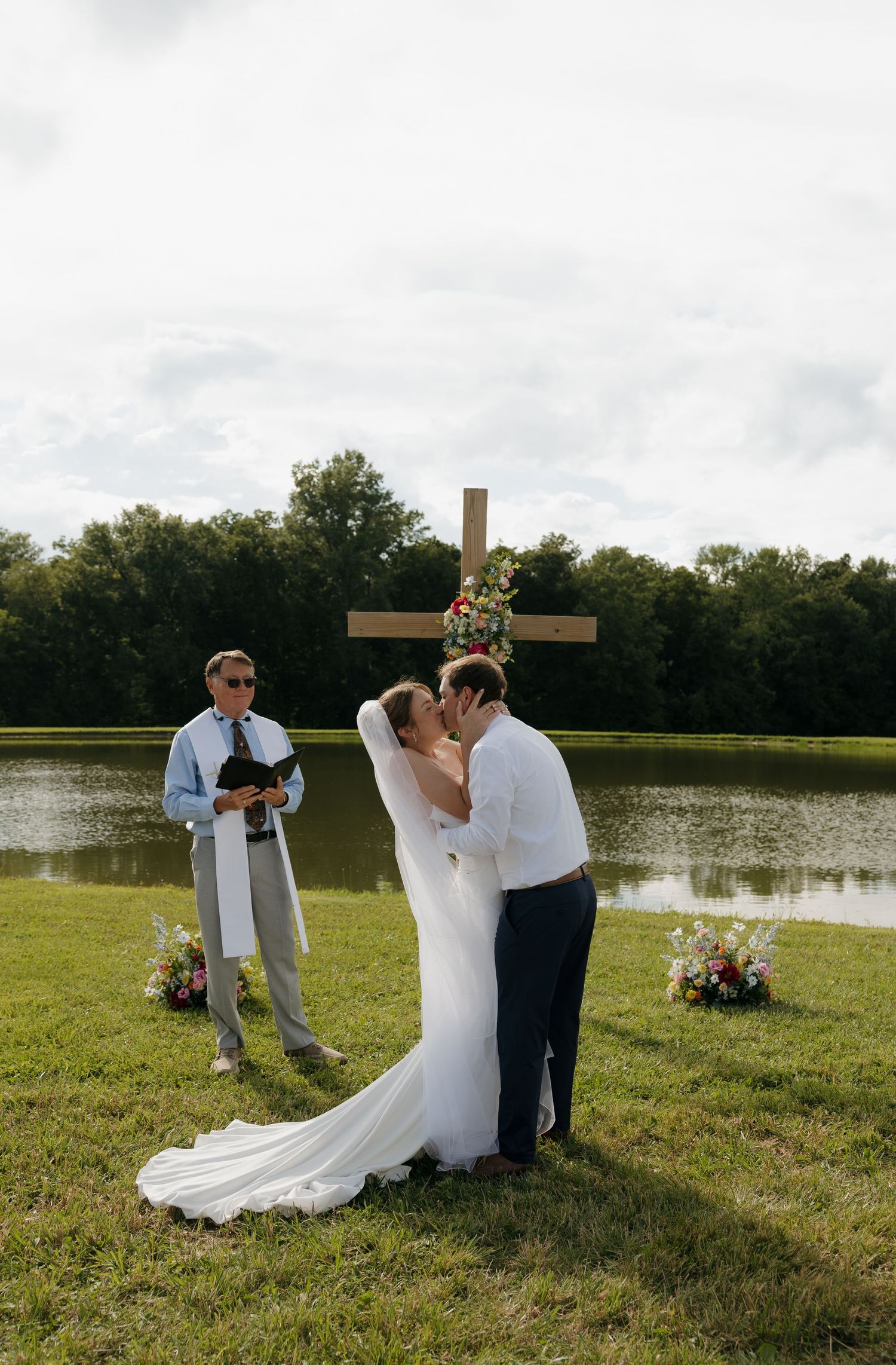 Bride and groom share first kiss at outdoor wedding ceremony by a lake with a floral cross altar.