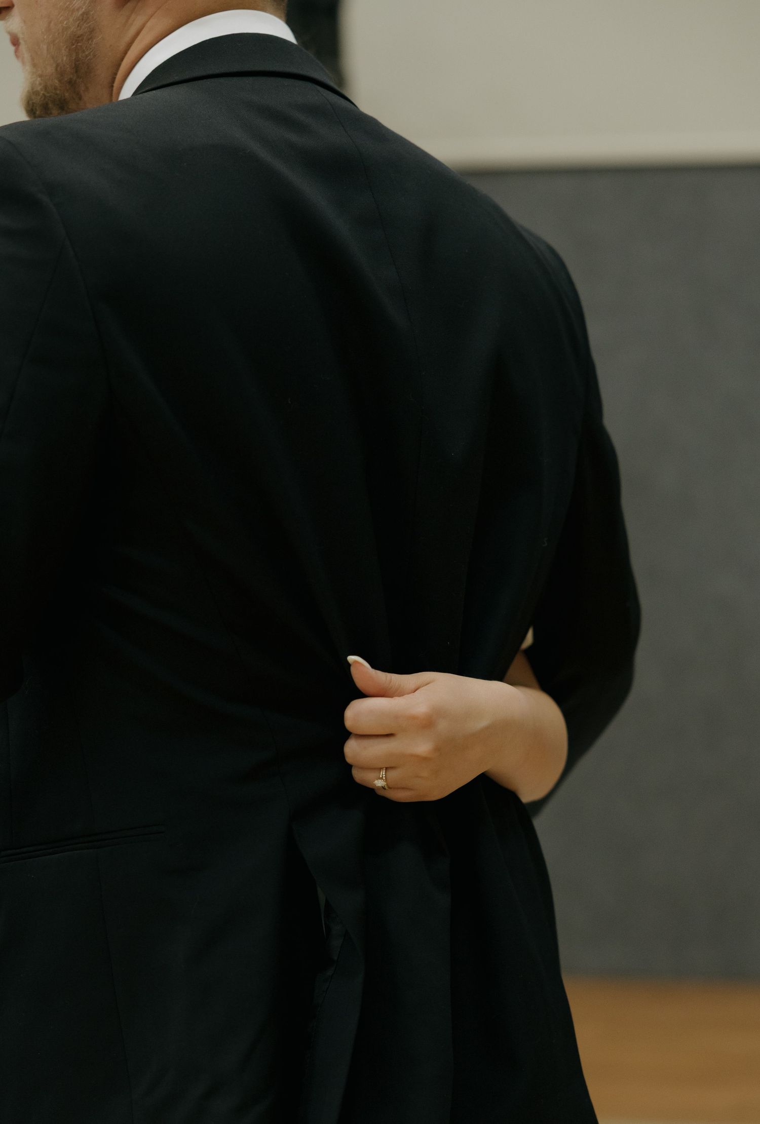A woman's hand with a ring gently grips the back of a man's black suit jacket during an embrace.
