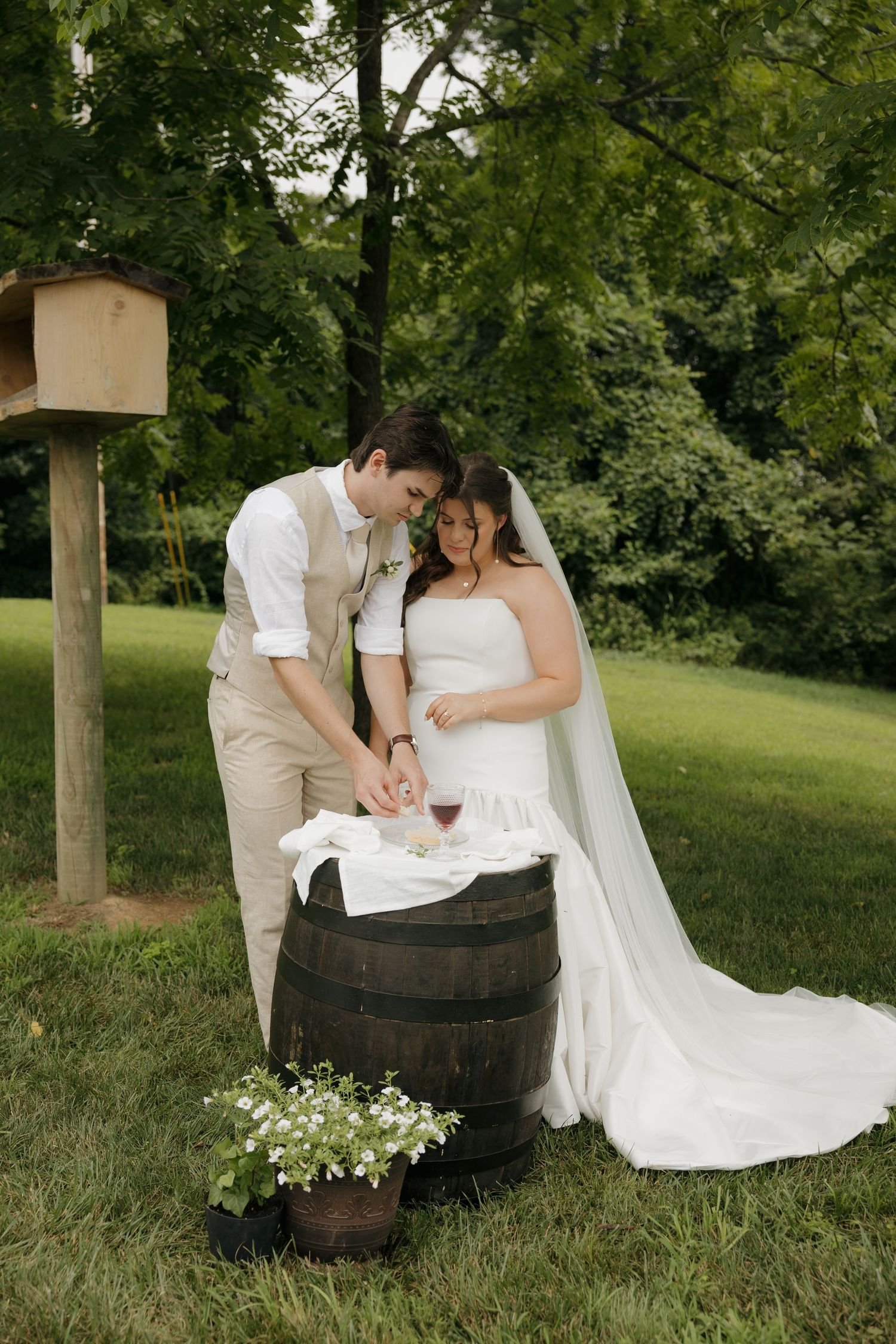 Bride and groom cutting cake on a rustic barrel outdoors, surrounded by lush greenery and white floral arrangements.