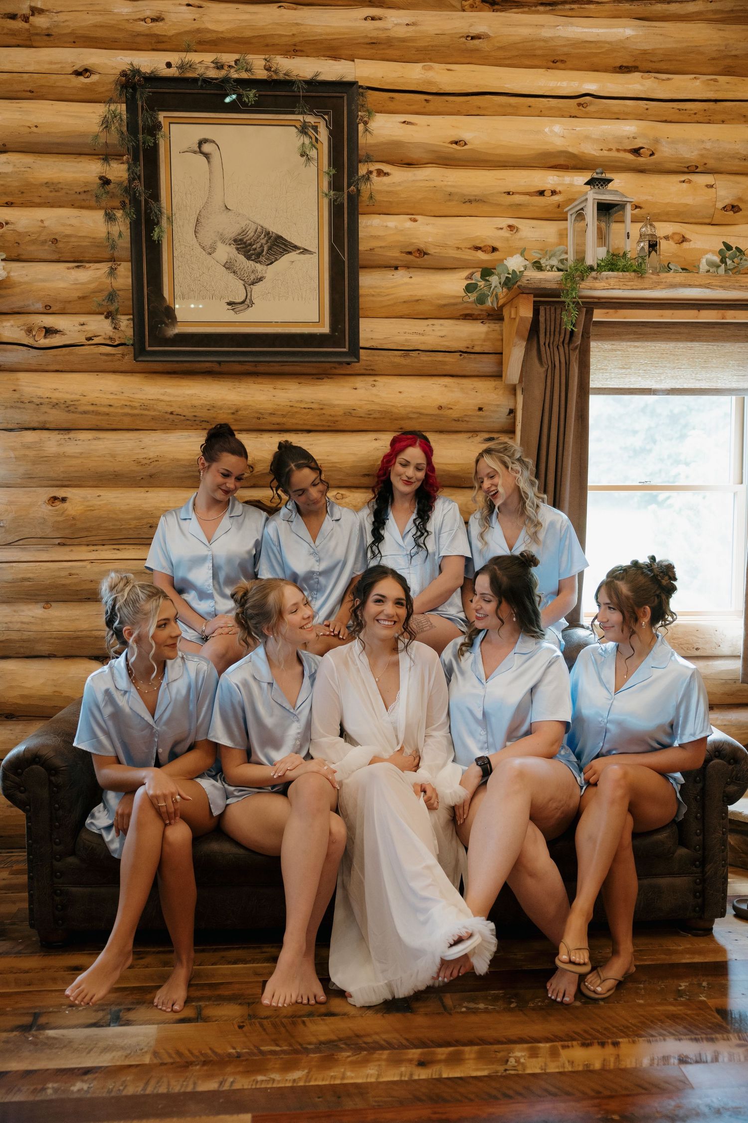 Bride in white robe surrounded by bridesmaids in light blue satin pajamas sitting in a rustic log cabin setting.