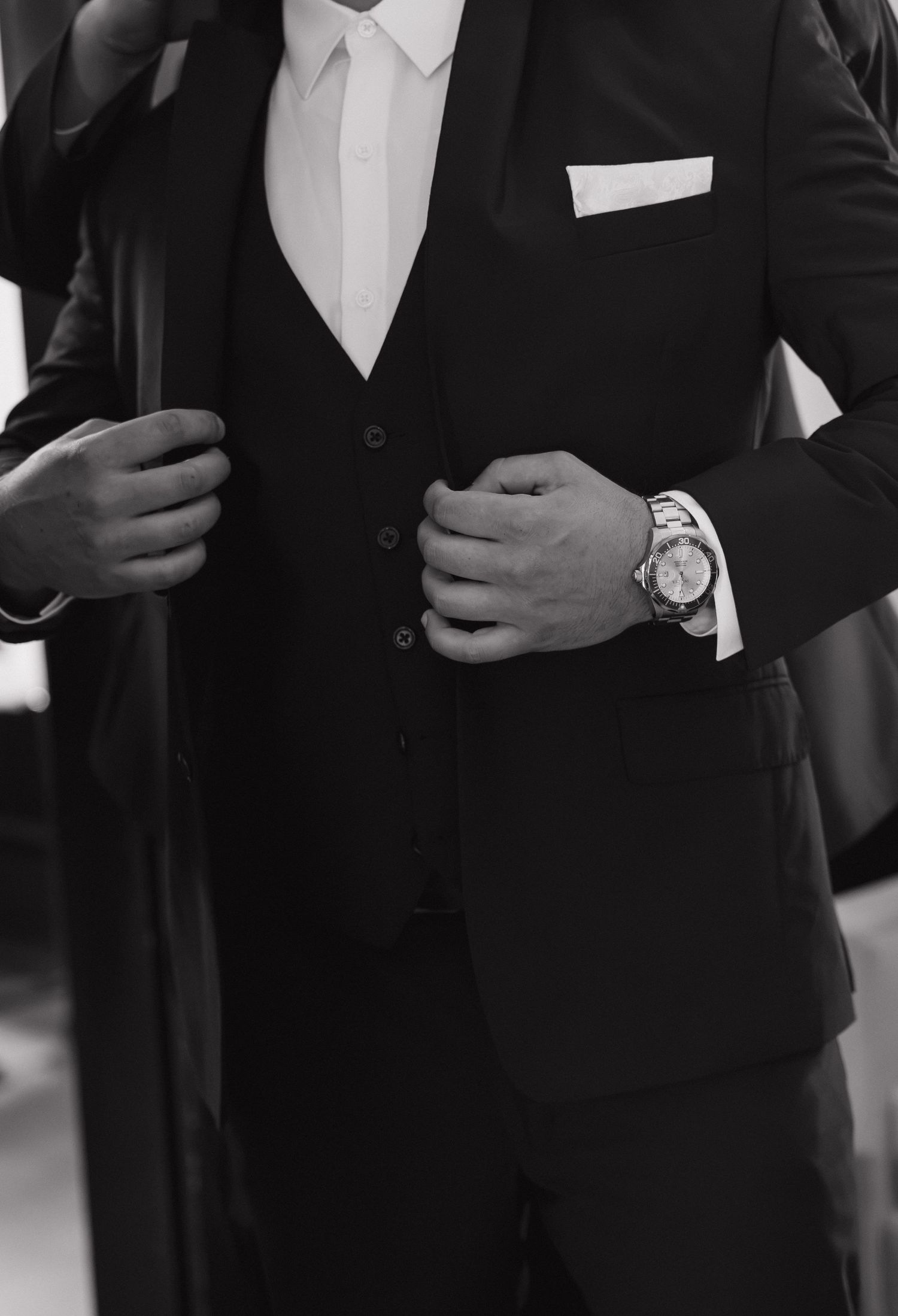 Man in black three-piece suit with white pocket square and luxury watch, elegant formal attire in black and white photo.