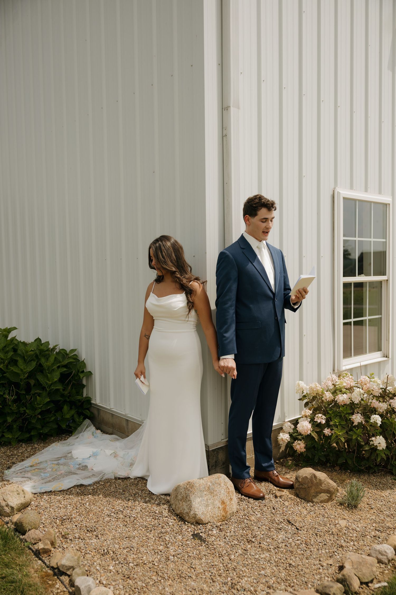 Bride and groom stand back-to-back outside a white barn, reading vows before their first look on their wedding day.