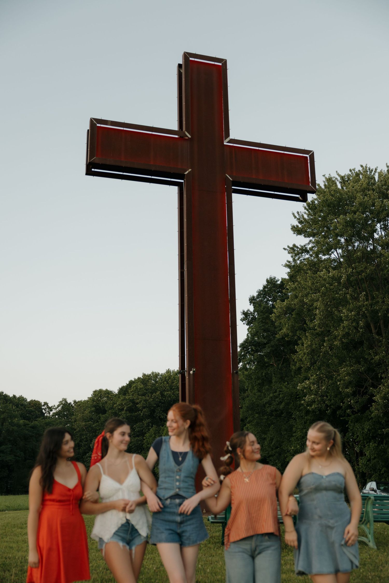 Five young women walk together in front of a large red Christian cross outdoors at dusk.