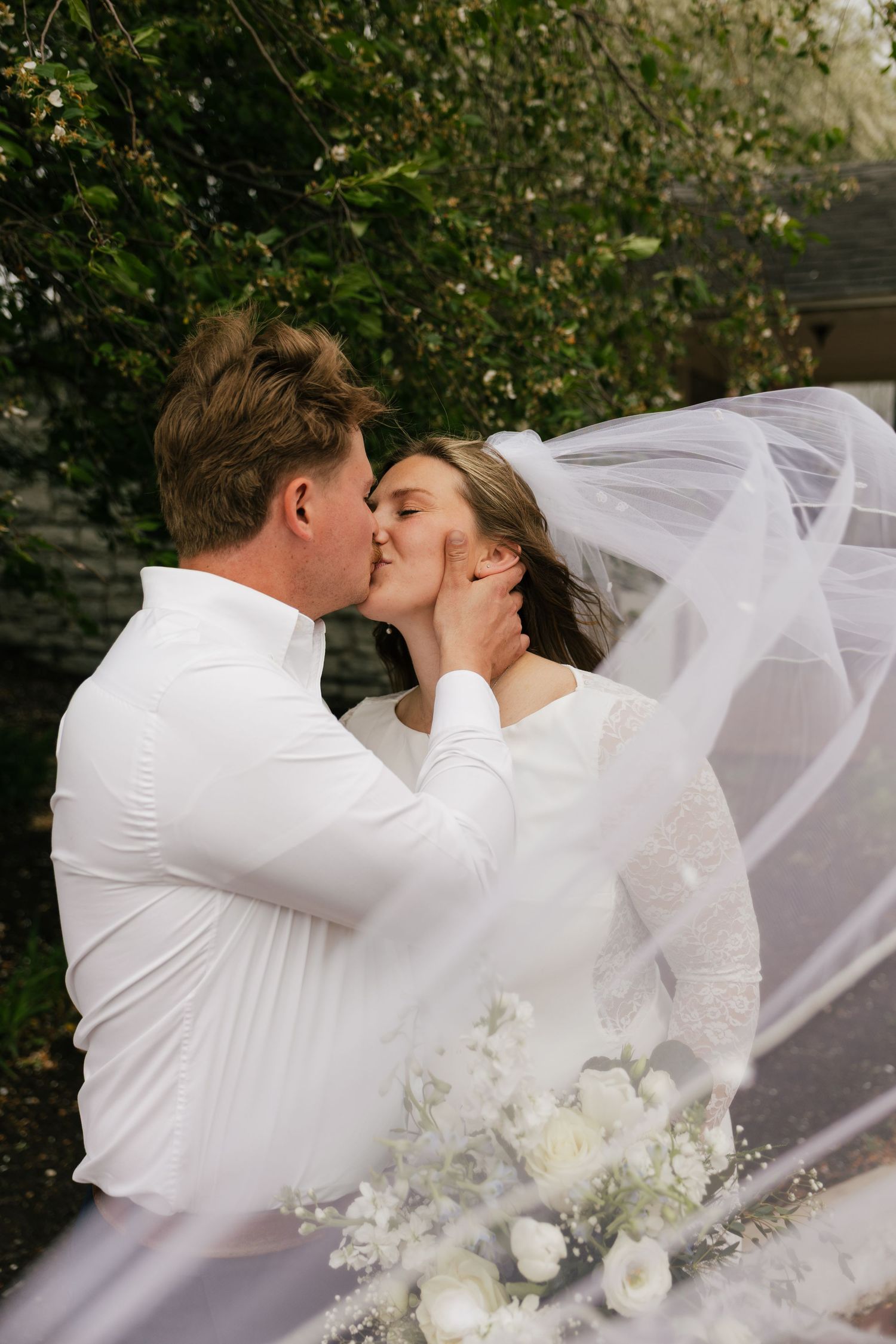 Bride and groom share a romantic kiss outdoors, with her white veil flowing in the wind, surrounded by lush green trees.