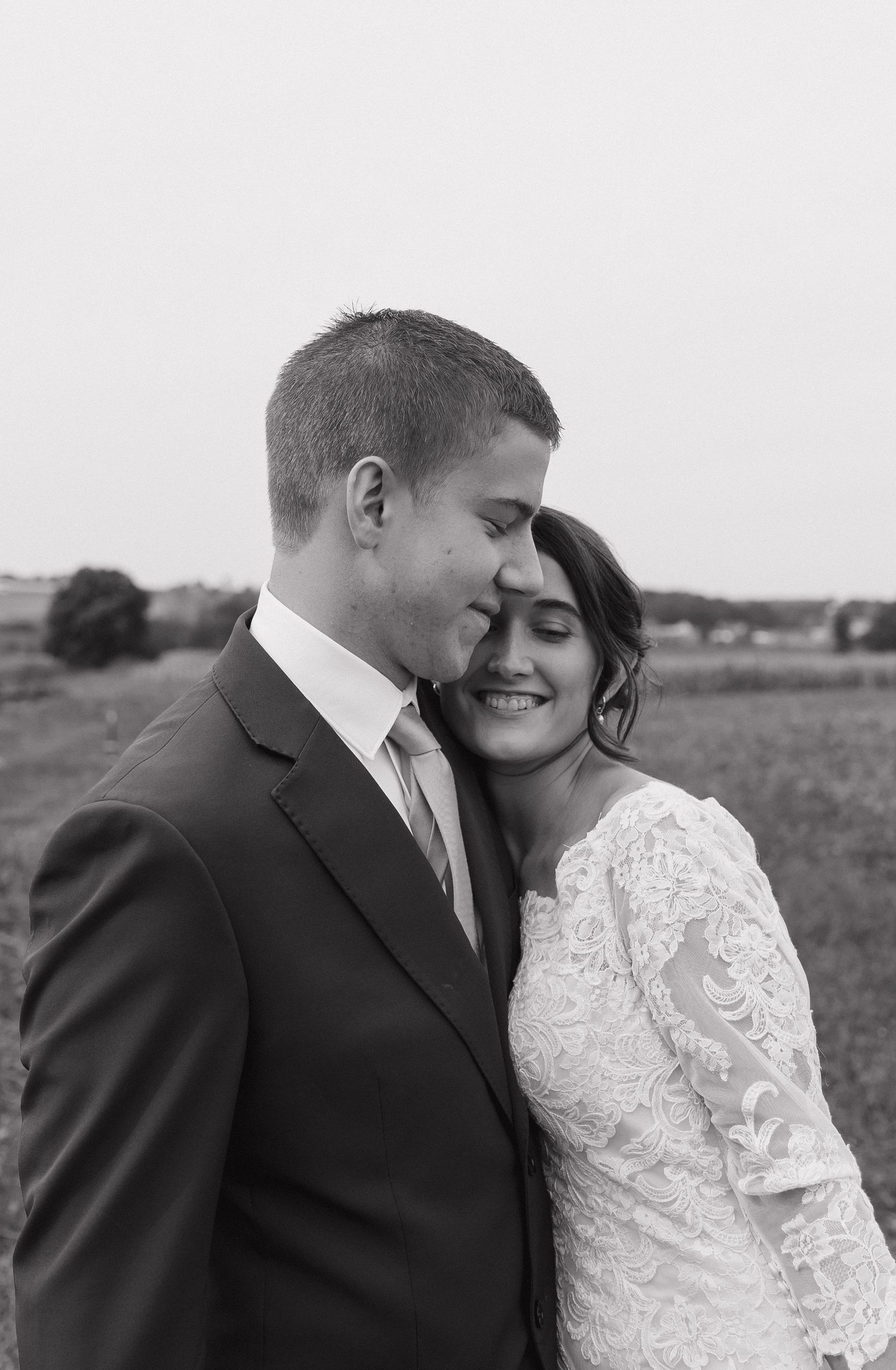Black and white wedding portrait of a smiling bride in a lace gown nestled against a groom in a dark suit in an open field.