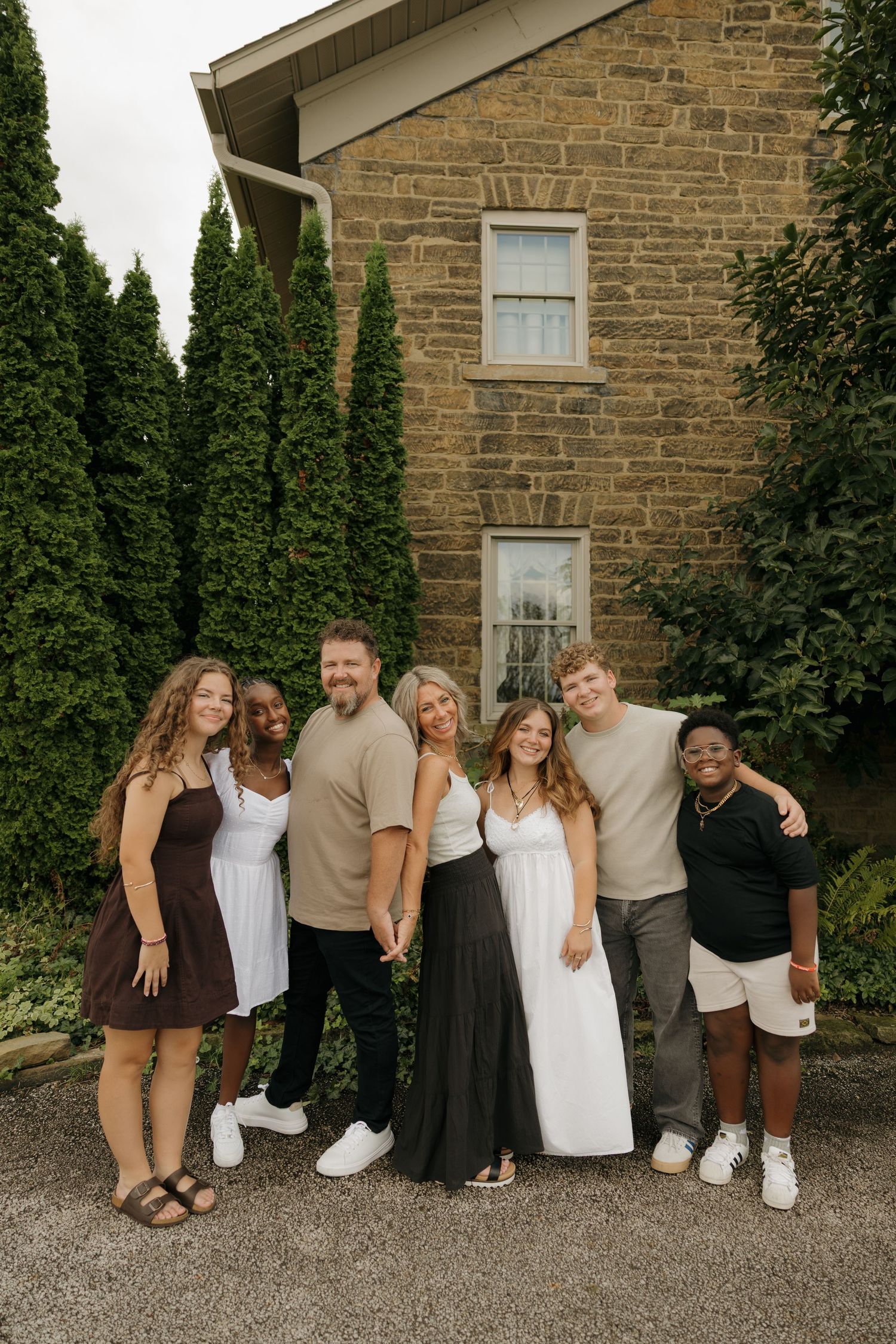A joyful family of seven poses together outdoors in front of a stone building surrounded by tall evergreen trees.