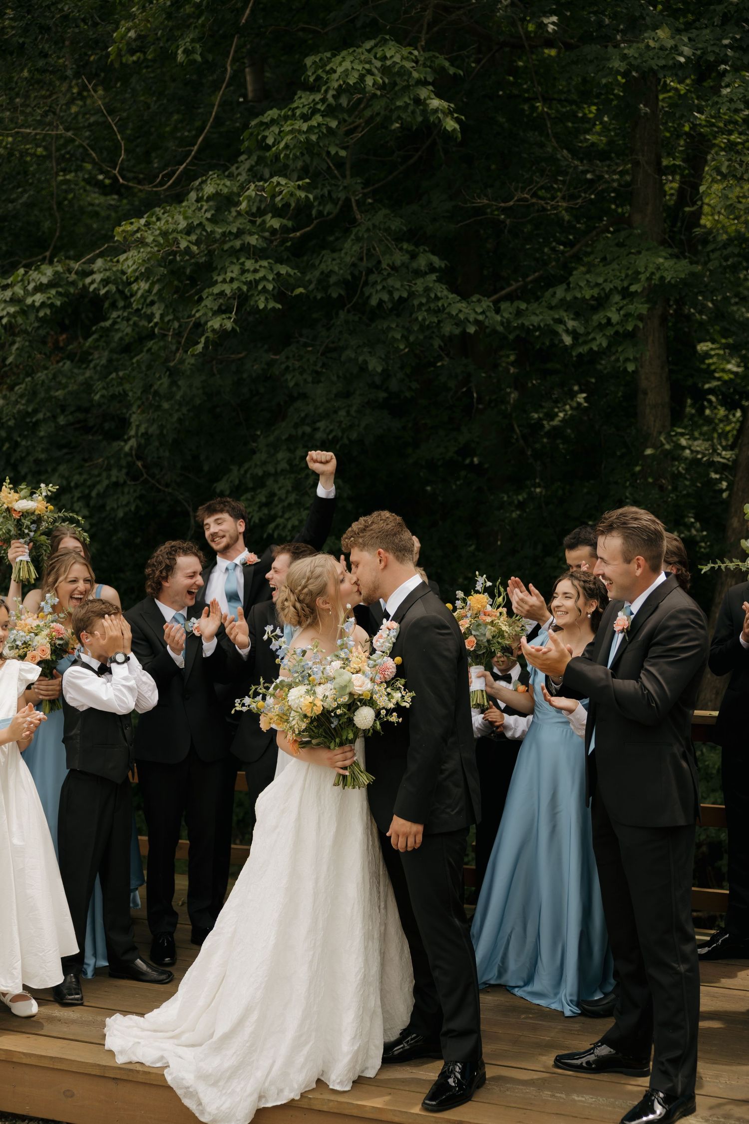 Bride and groom share a kiss surrounded by cheering wedding party outdoors with lush green trees in background.