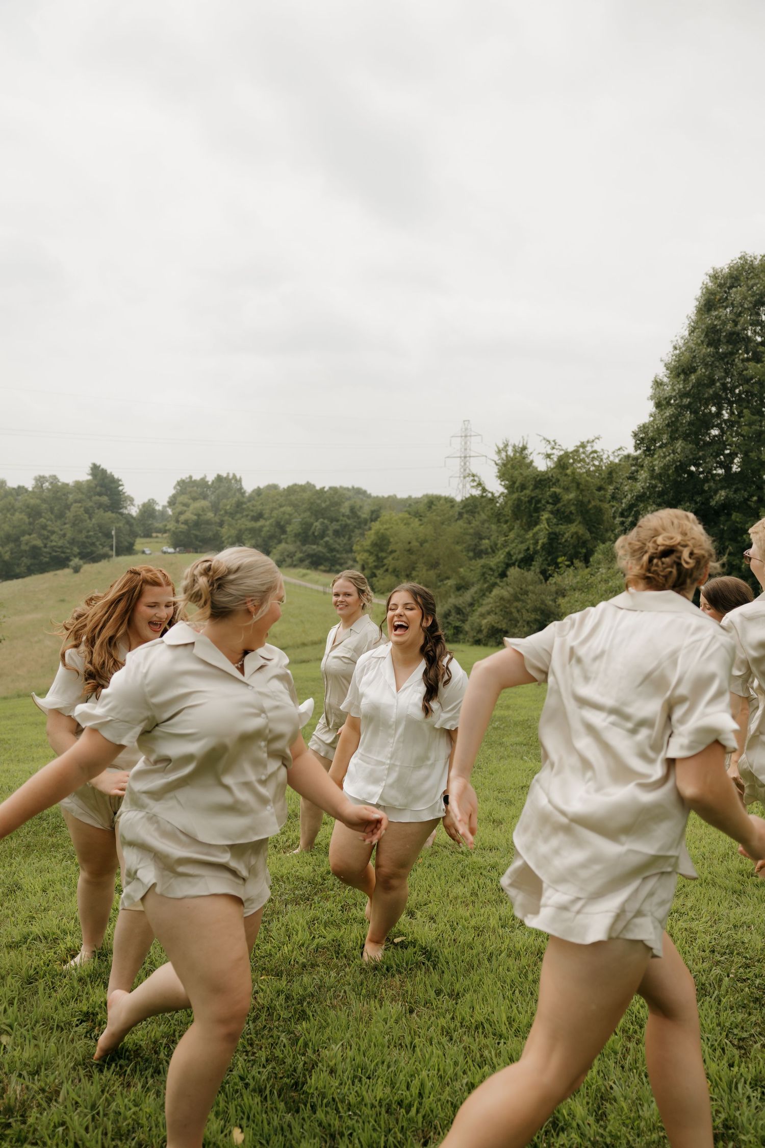 Group of women in matching white outfits laughing and running together on a lush green hillside on a cloudy day.