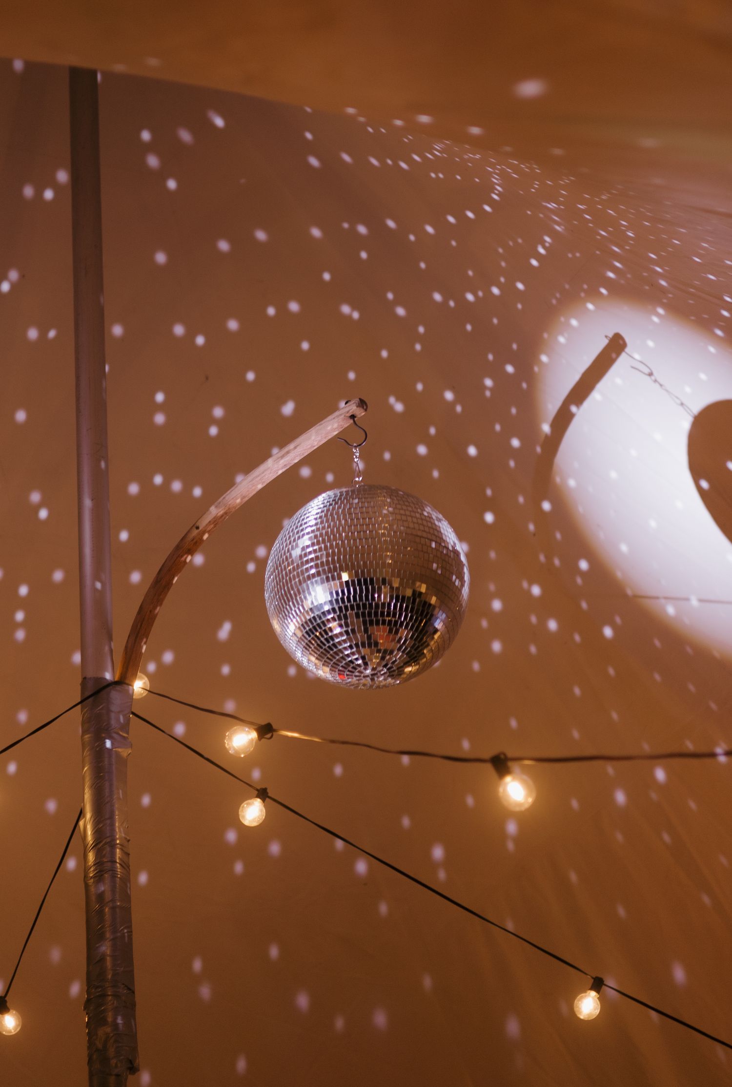 A shimmering disco ball hangs from a curved rod, casting sparkling light dots on a warm amber wall with string lights below.