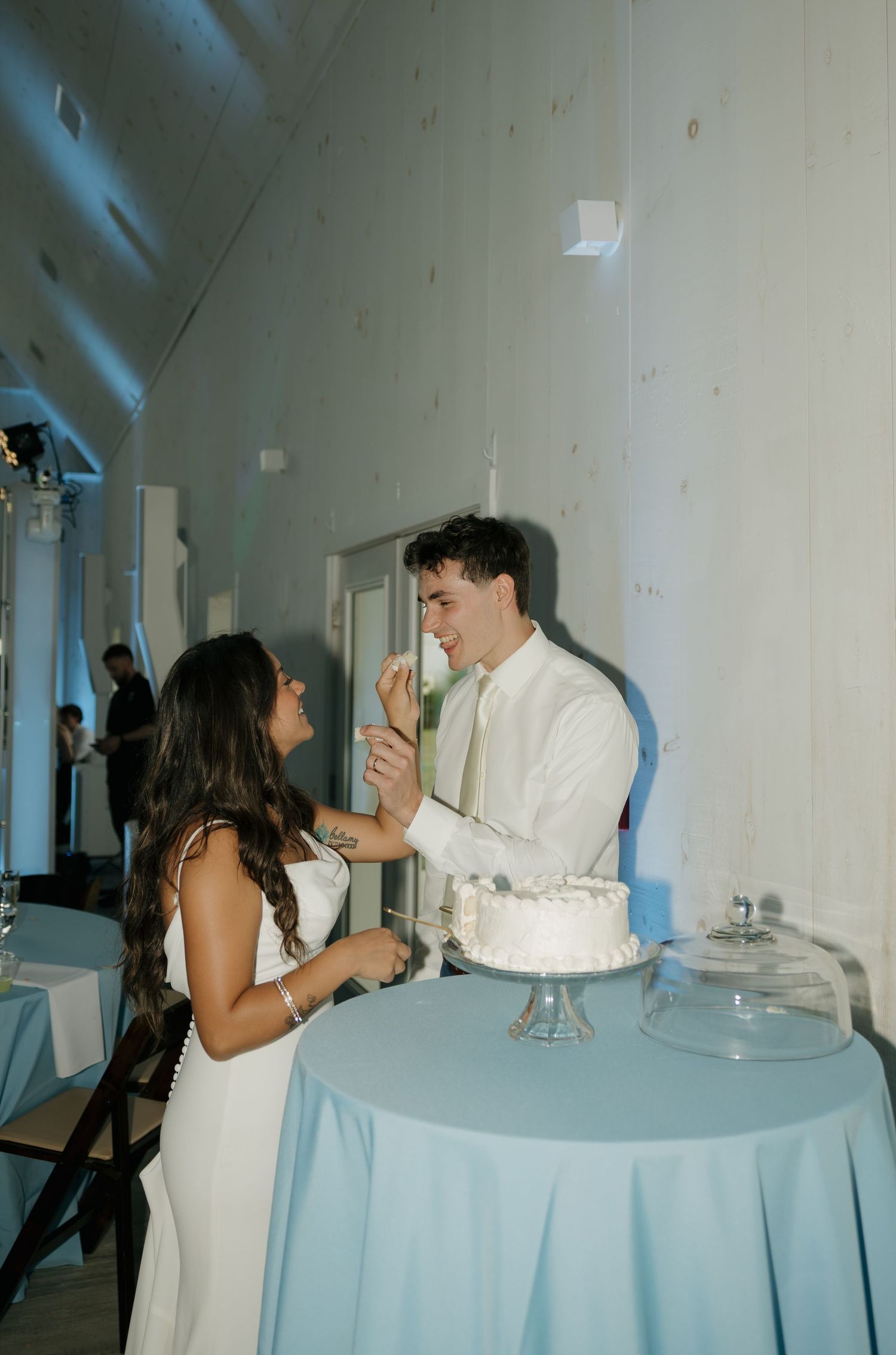 Bride feeds groom wedding cake at elegant blue-themed reception inside a white barn venue.