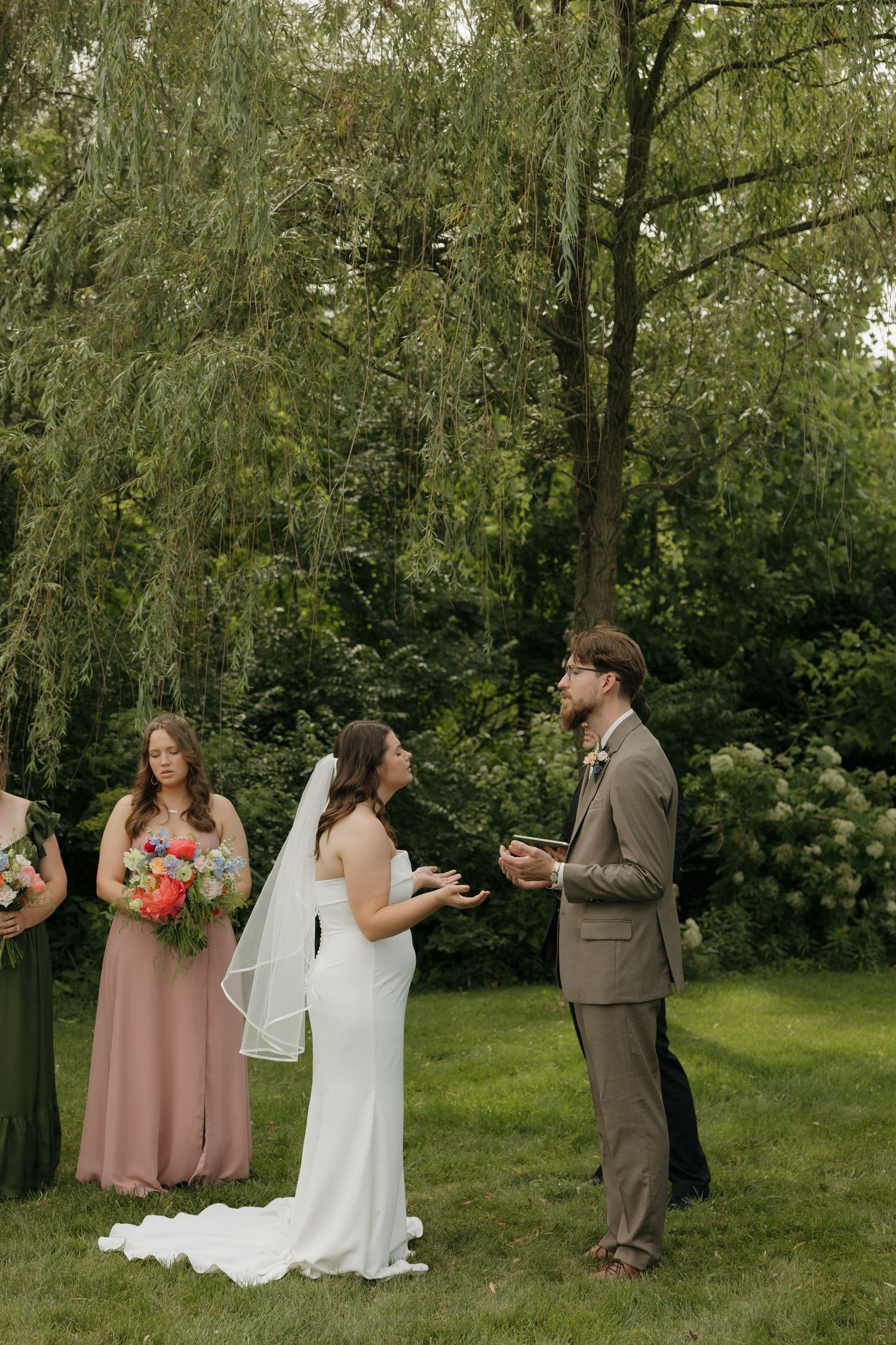 Bride and groom exchange vows outdoors under a weeping willow tree, with bridesmaids in pink and green dresses nearby.