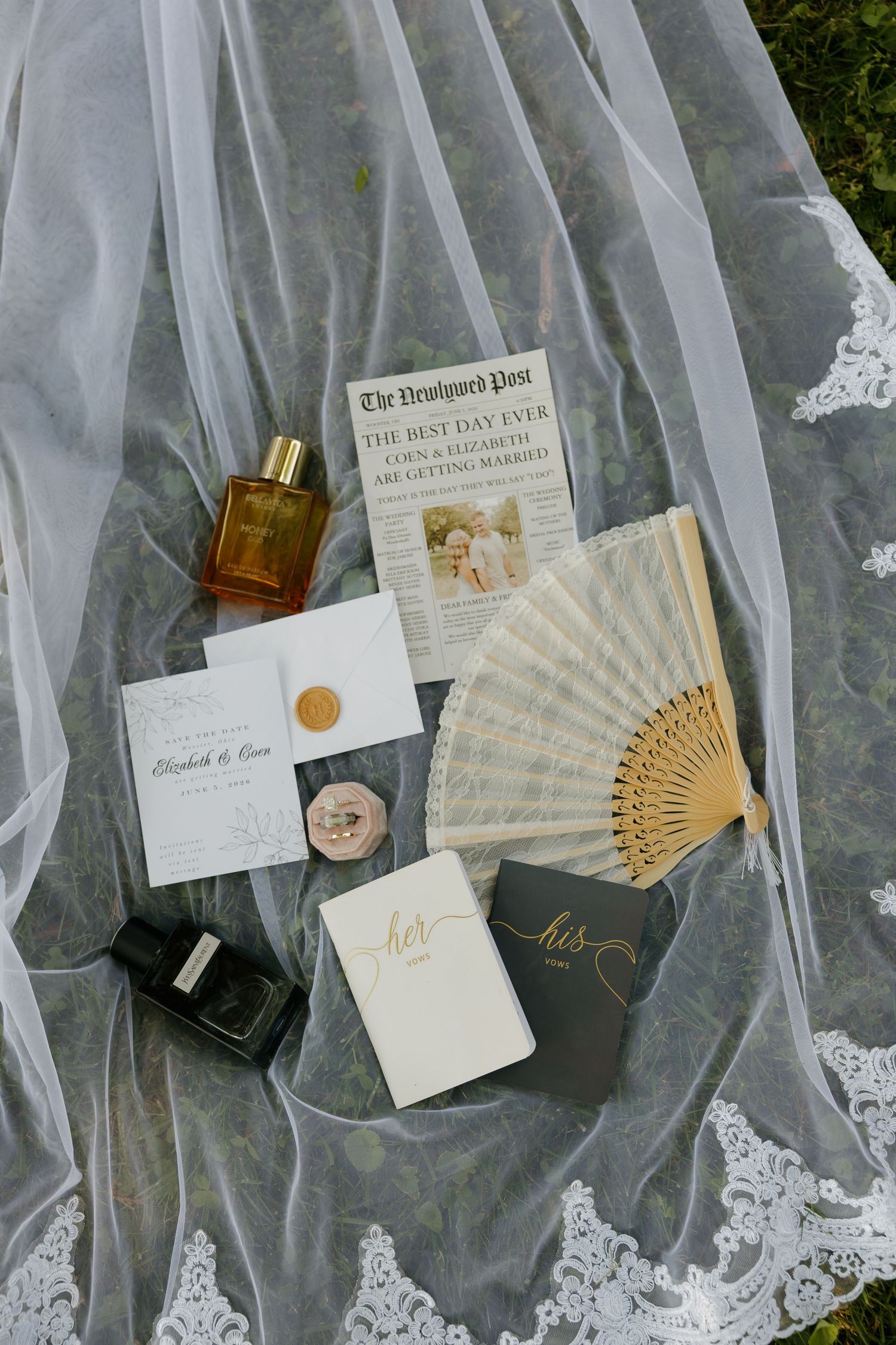 Elegant wedding flat lay featuring invitation suite, perfume, hand fan, vow books, and lace veil on grass.
