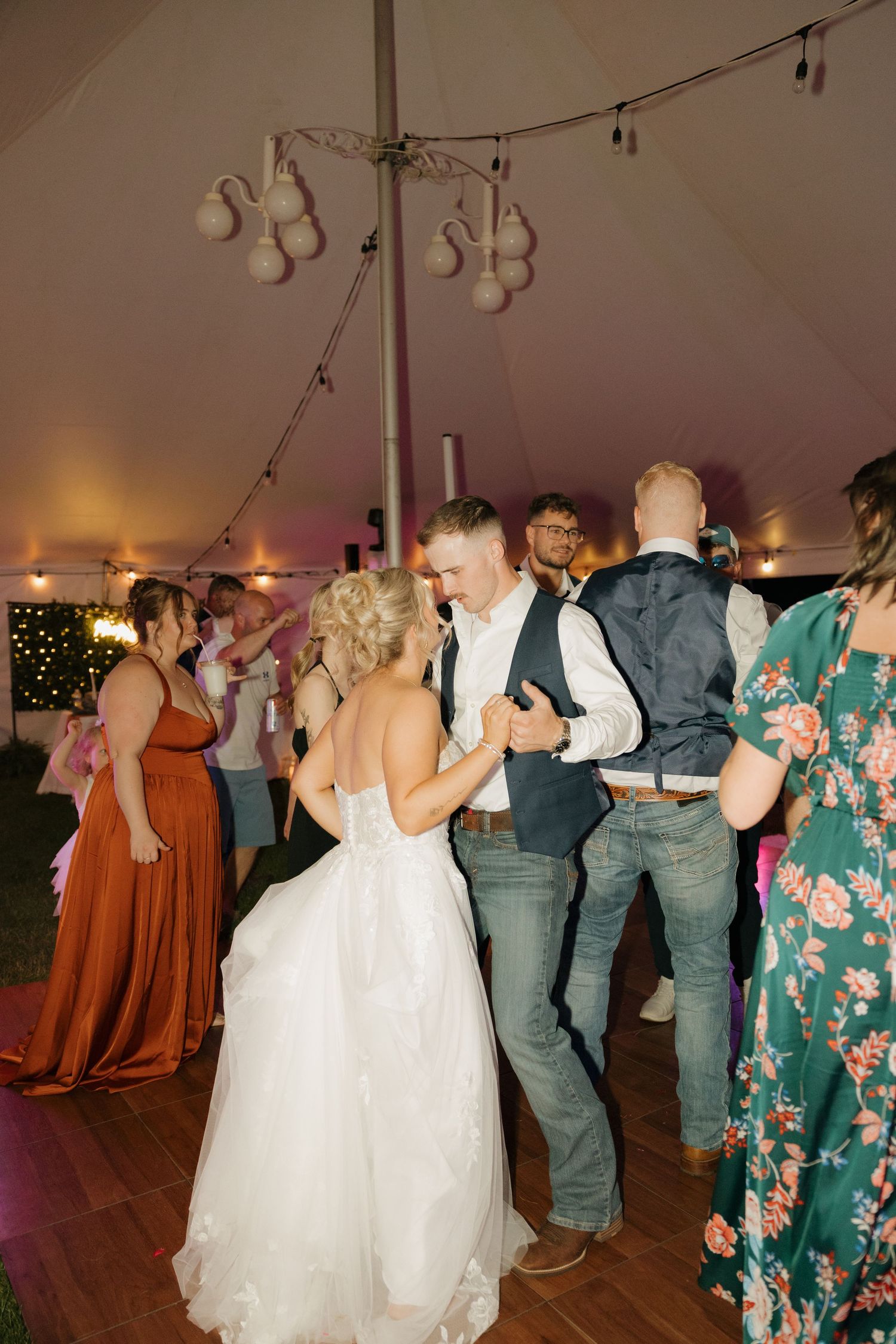 Bride in white gown dancing closely with groom in navy vest surrounded by wedding guests under tent.