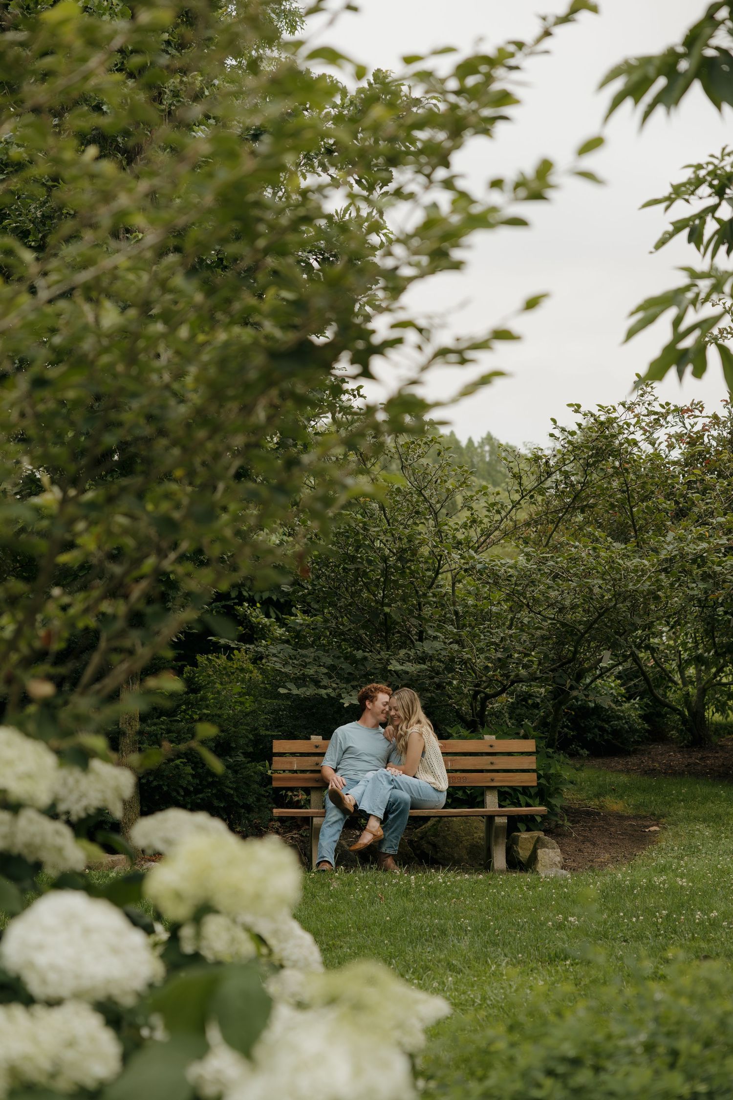 Couple sitting close together on a wooden bench surrounded by lush green trees and white hydrangeas in a garden.