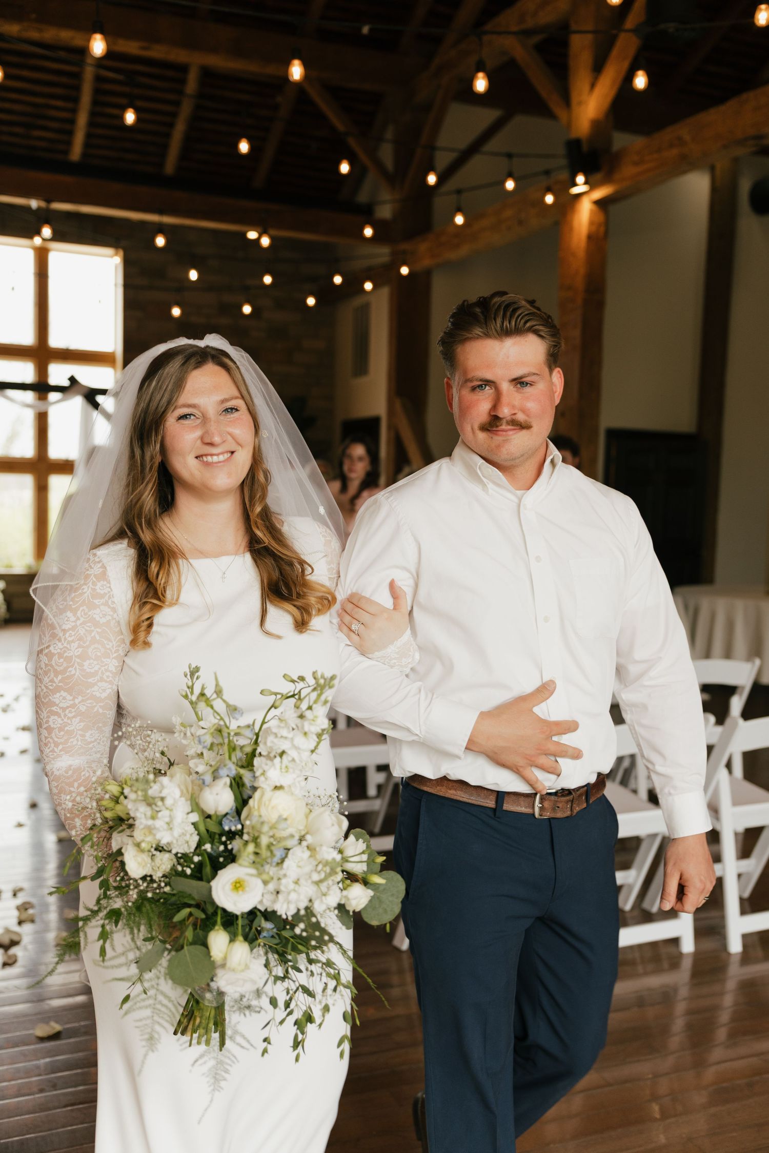 Bride in white lace gown holding floral bouquet walks with groom in white shirt and navy pants at rustic barn wedding.