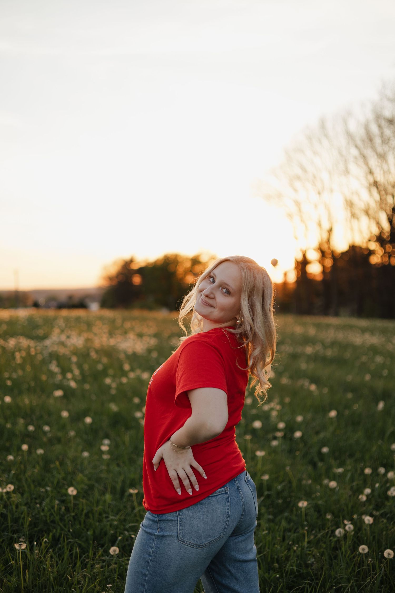 Young blonde woman in red t-shirt and jeans posing in a dandelion field during golden hour sunset.