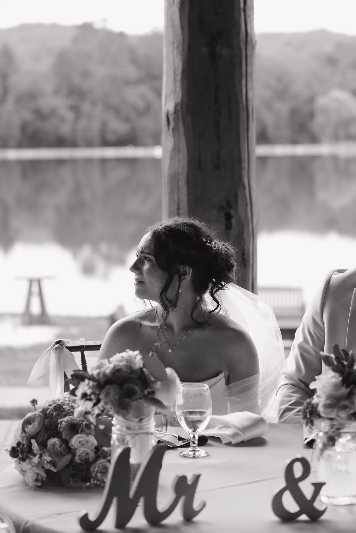 Black and white photo of a bride seated at a wedding reception table with Mr & signs and floral arrangements.