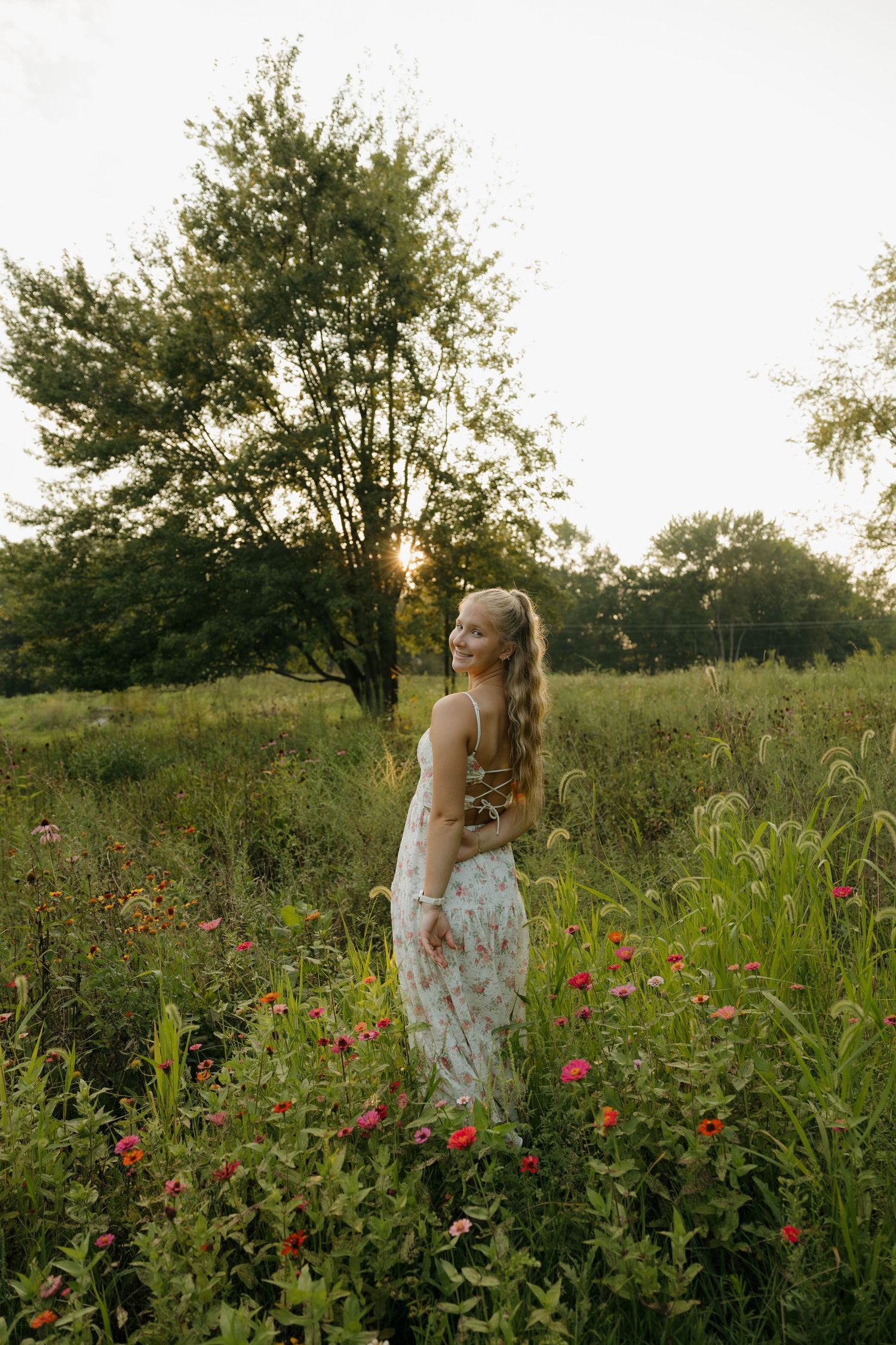 Young woman in floral dress standing in wildflower field at golden hour sunset with lush green trees behind her.