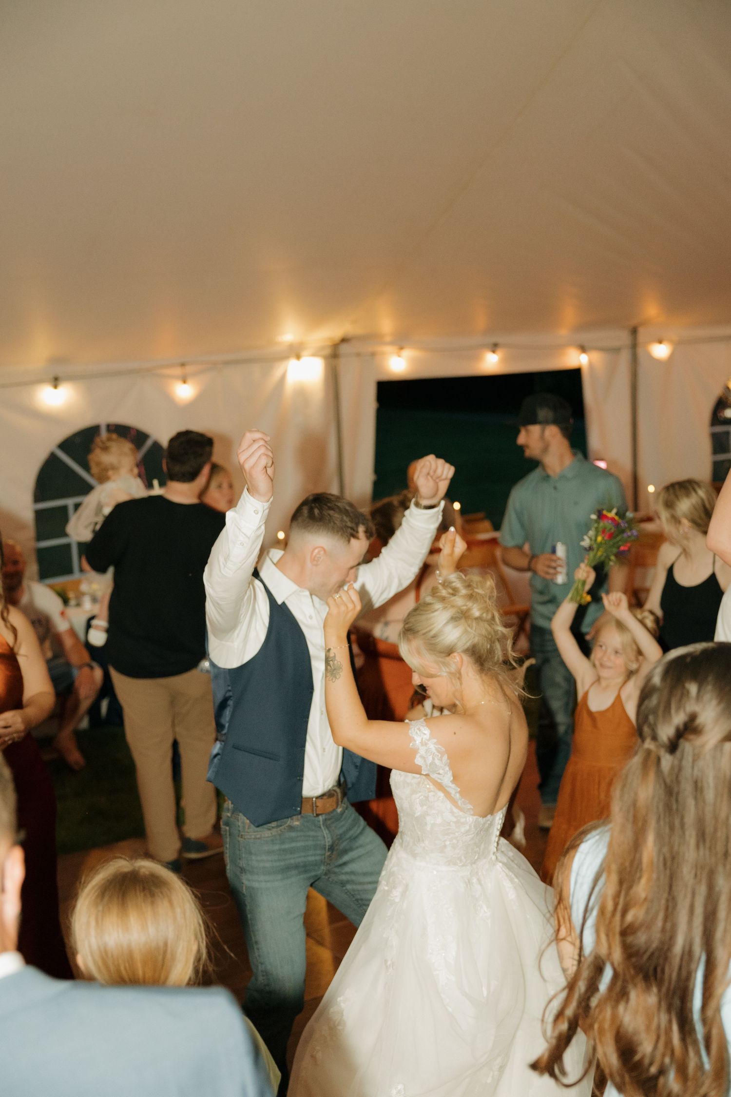Bride and groom dancing together joyfully surrounded by guests on lit wedding reception dance floor.