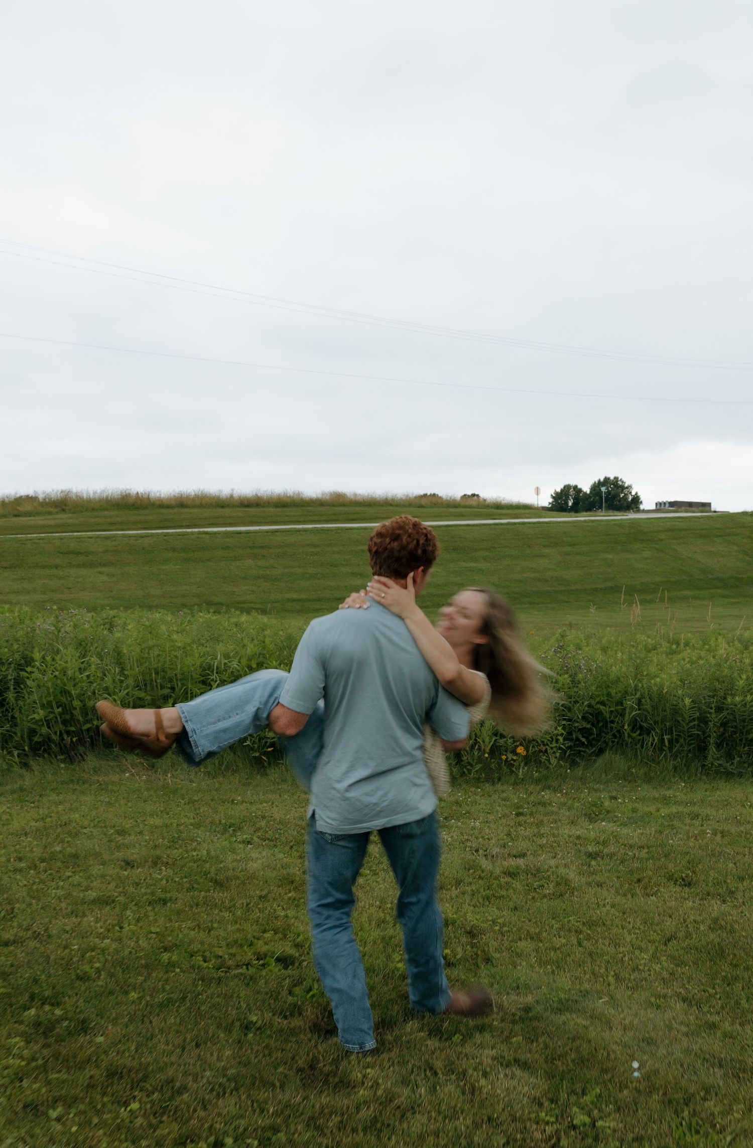 A man carries a woman in his arms while spinning in a lush green field on a cloudy day.