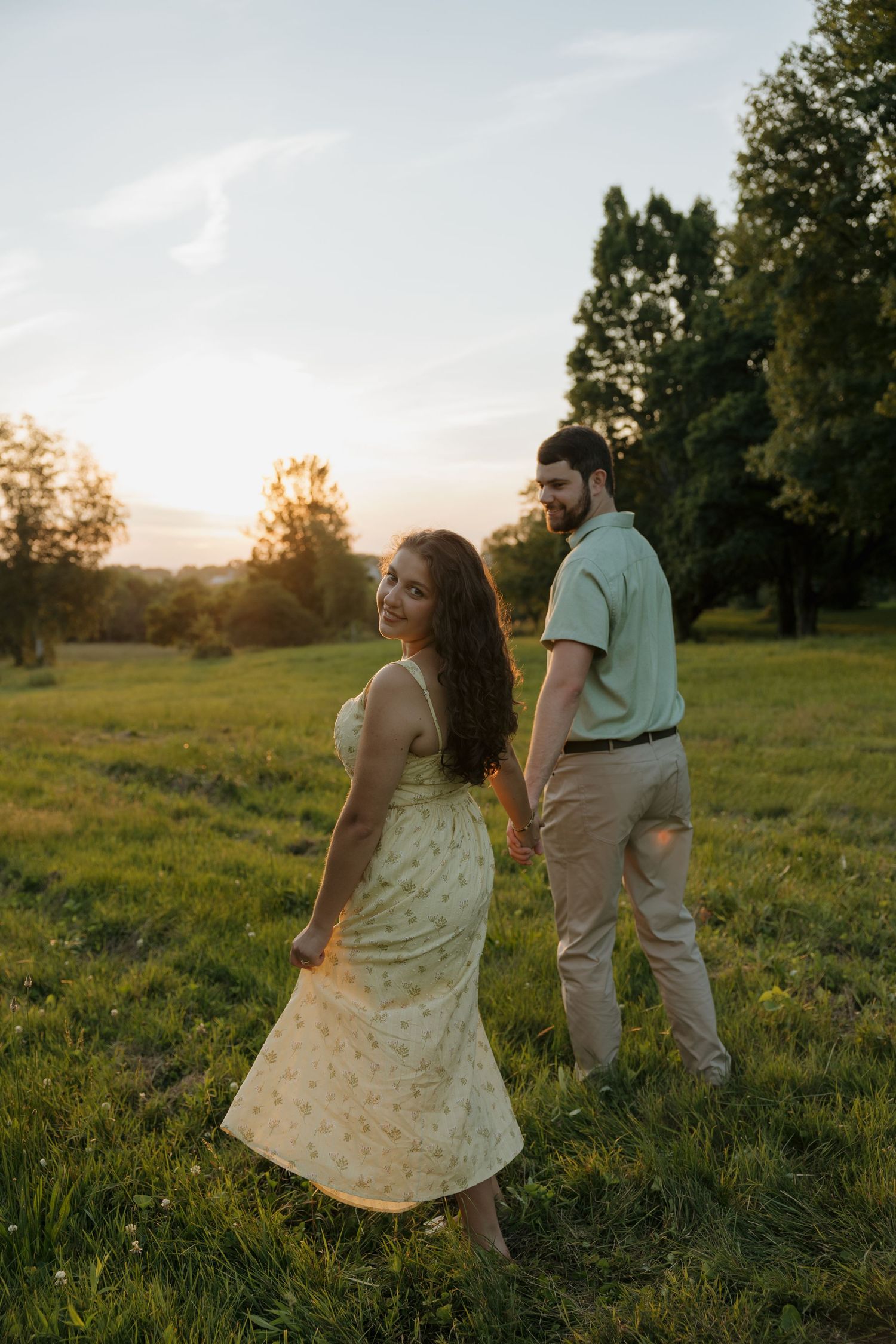 Couple holding hands in a sunlit green field at golden hour, woman in floral dress smiling back at camera.