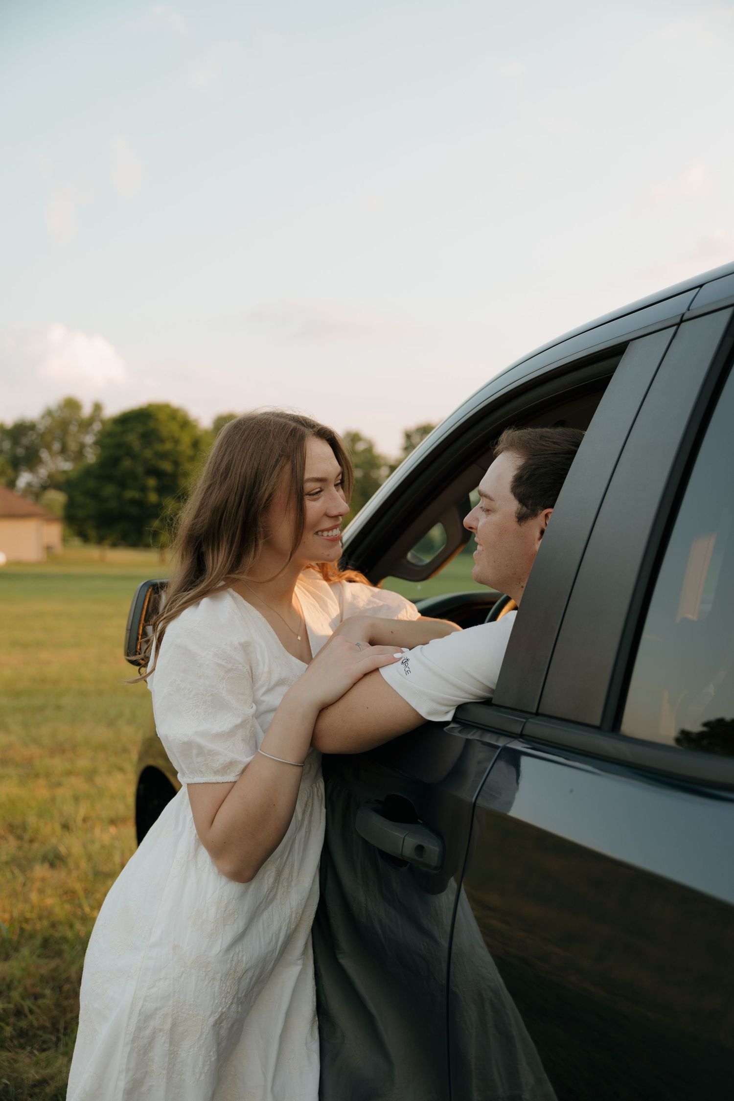 Smiling bride in white dress holding hands with groom through car window in a sunny green field.