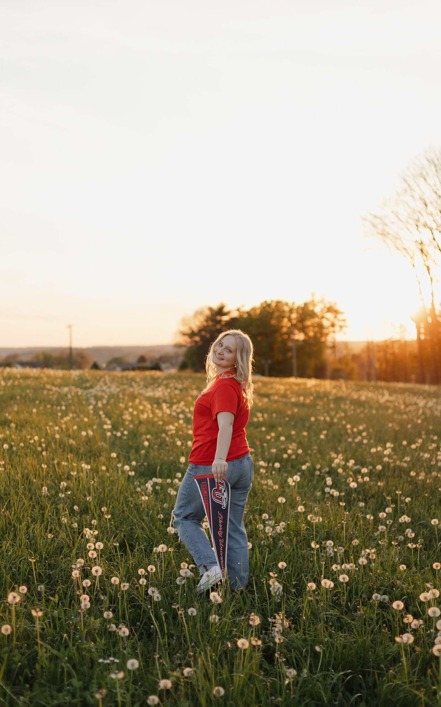 Blonde woman in red shirt stands in a sunlit dandelion field at sunset holding a pennant.