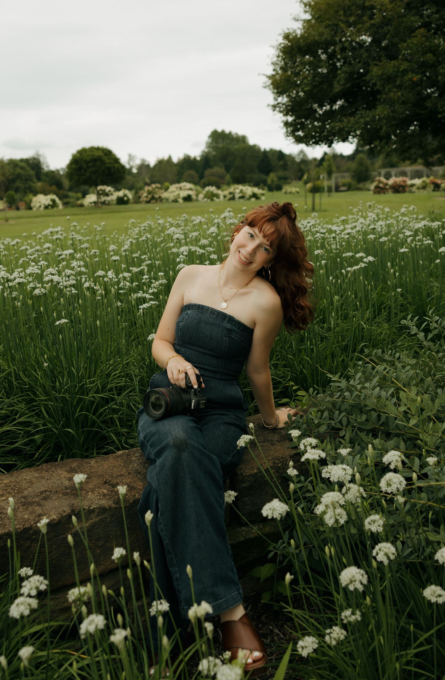 A red-haired woman in a denim jumpsuit sits among white wildflowers in a lush green garden, holding a camera.