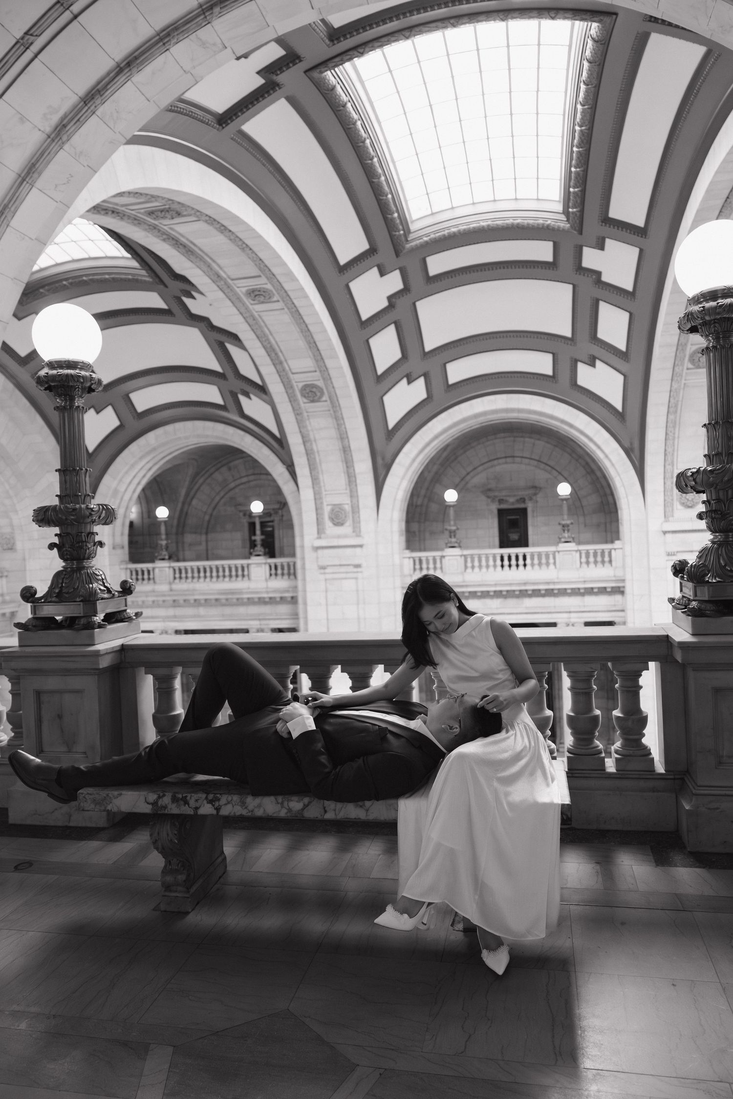 Black and white photo of a couple posing in an elegant arched hall, woman in white dress sits beside man lying on bench.