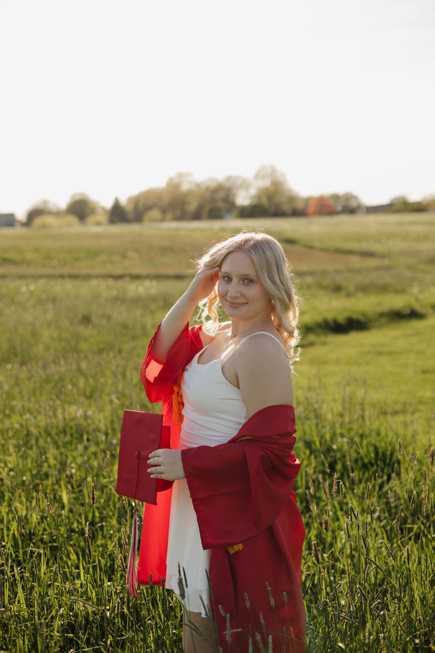 Blonde woman in red graduation gown holding cap in sunny green field at golden hour.