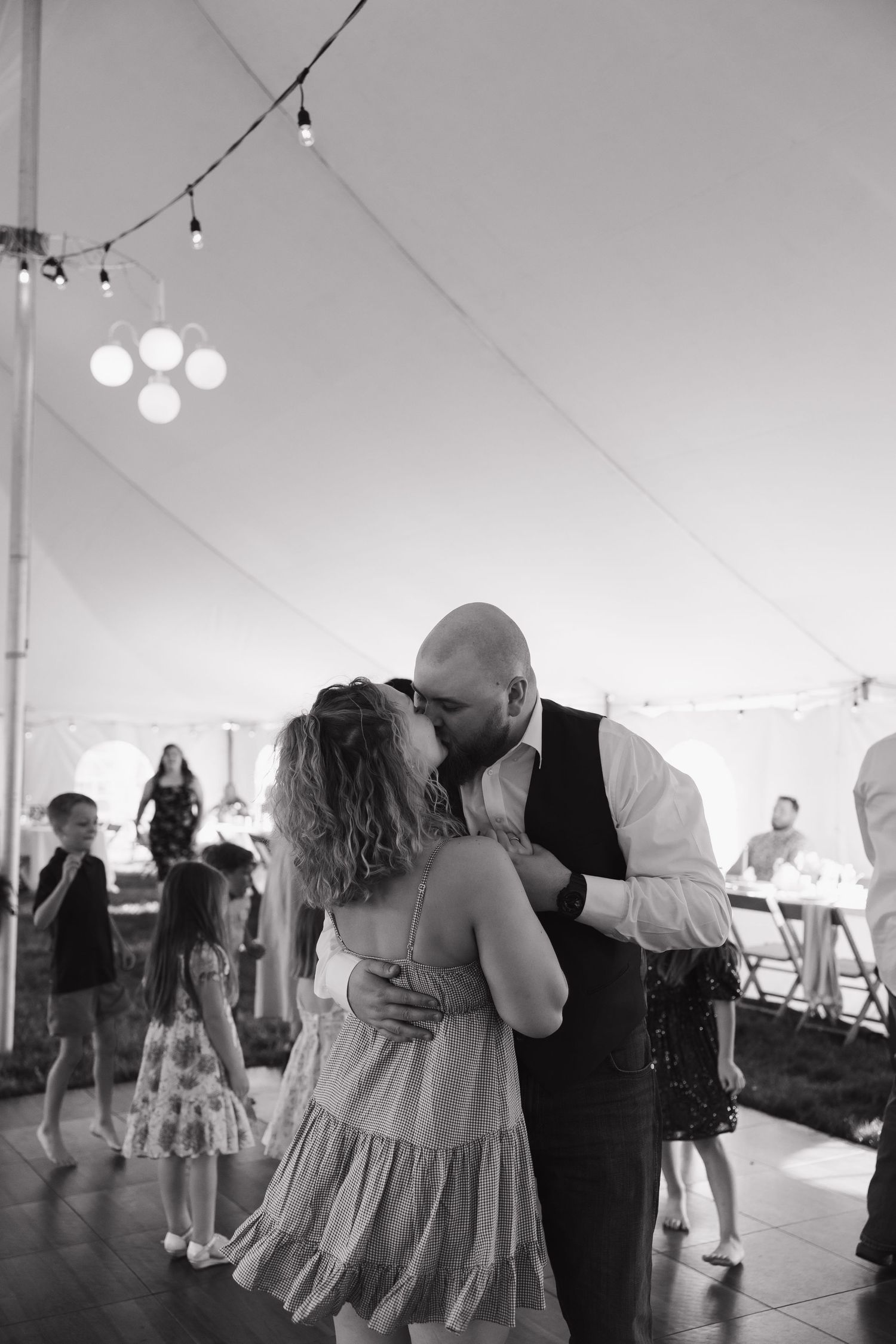 Black and white photo of couple sharing a romantic kiss on the wedding reception dance floor under string lights.