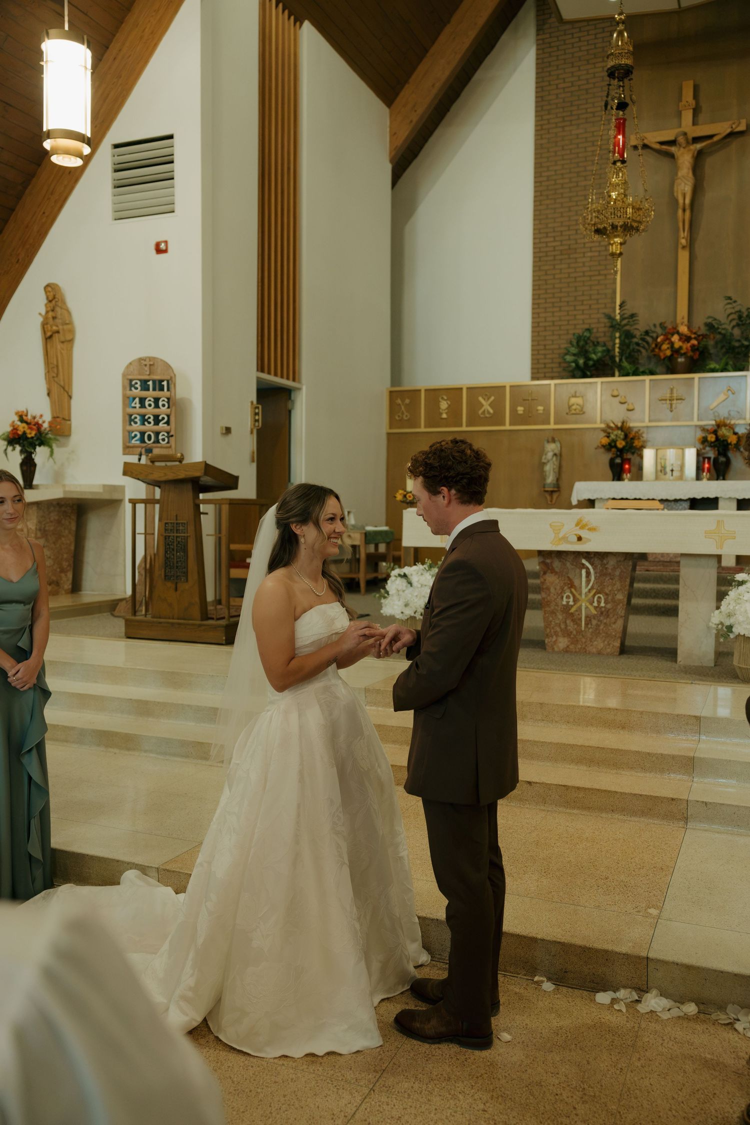 Bride in a white gown and groom in a brown suit exchange vows at the altar of a church, with a crucifix and bridesmaid nearby.