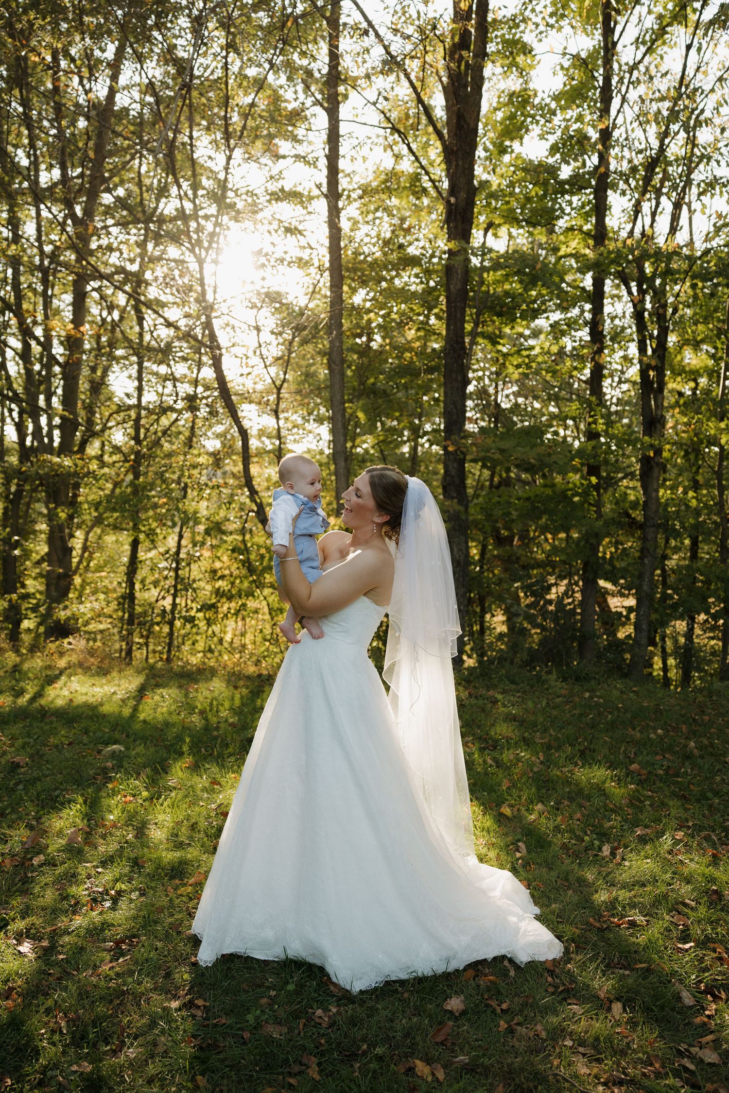 A bride in a white gown and long veil holds a baby in a sunlit forest clearing, with golden light filtering through tall trees.