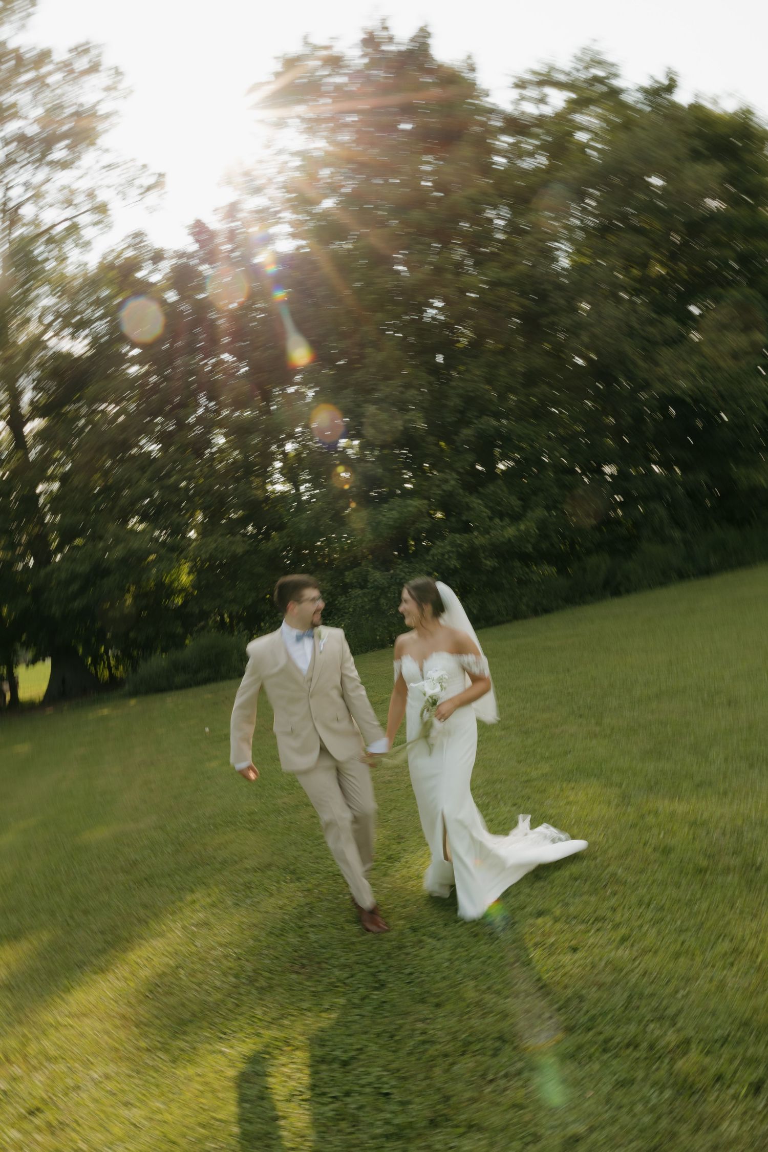Bride and groom walk hand-in-hand across lush green lawn in golden sunlight, bride in white gown, groom in beige suit.