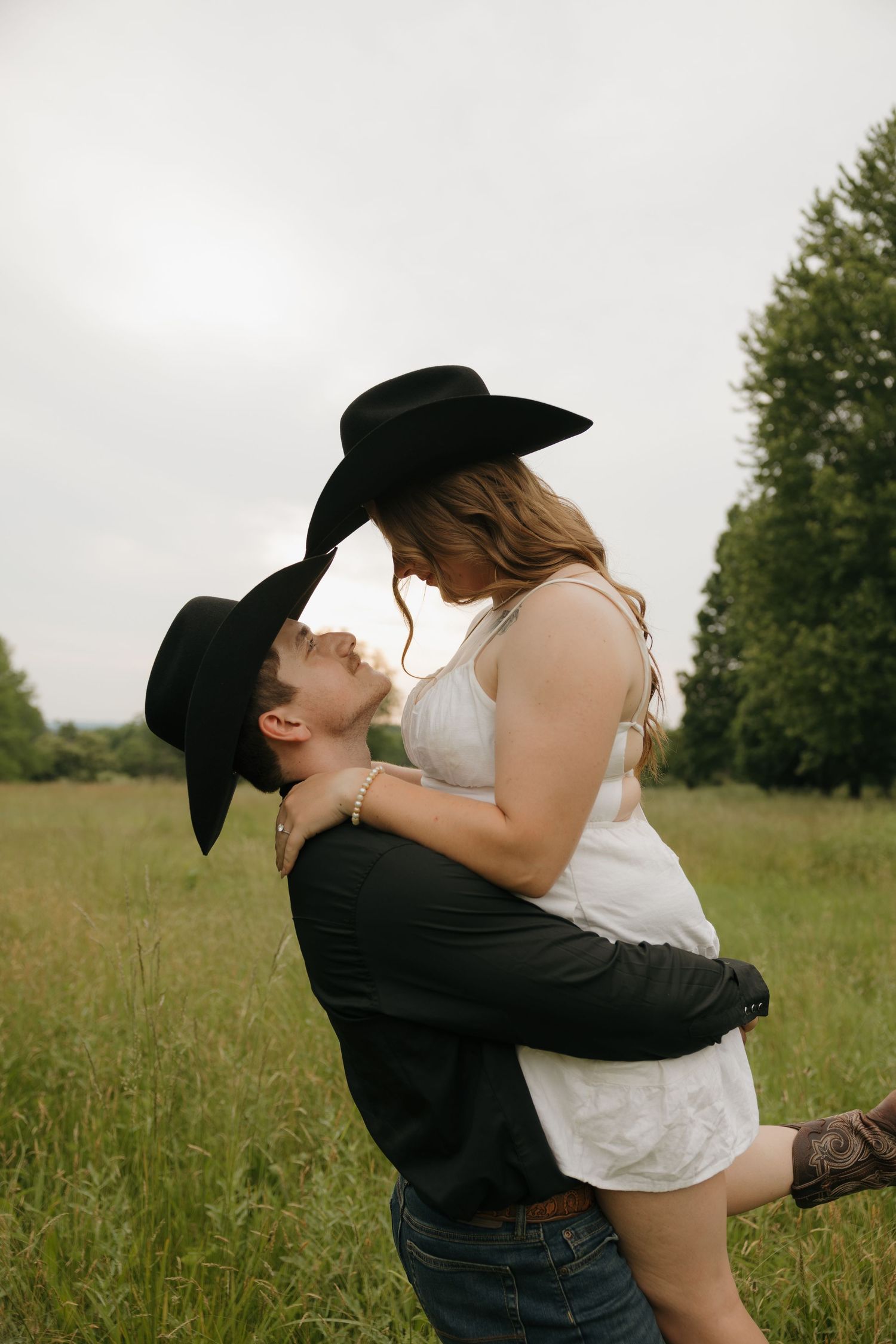 A couple in black cowboy hats share a romantic moment in a green field, as the man lifts the woman in a white dress.
