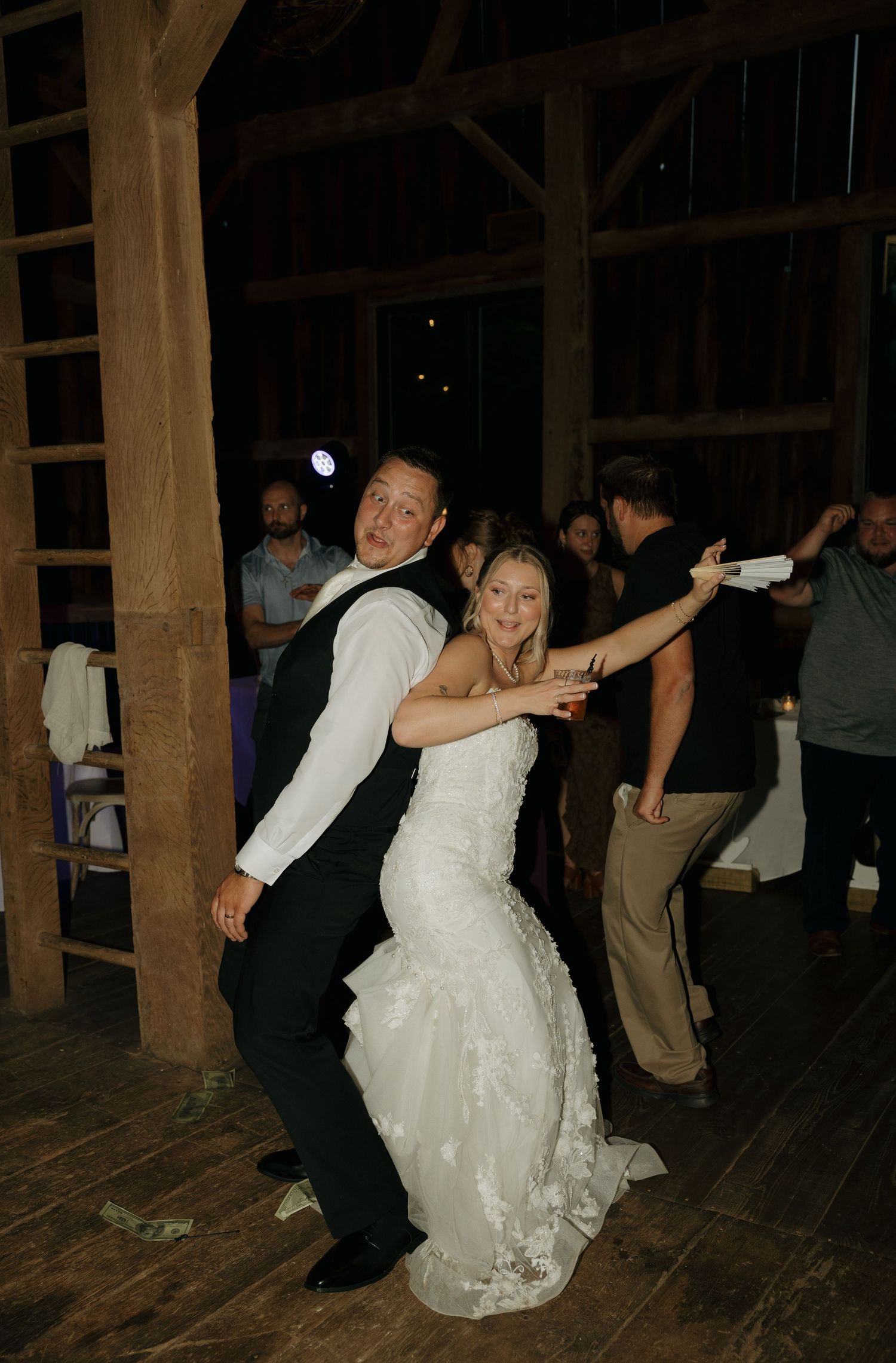 Bride and groom dancing joyfully at rustic barn wedding reception, surrounded by guests celebrating on wooden dance floor.