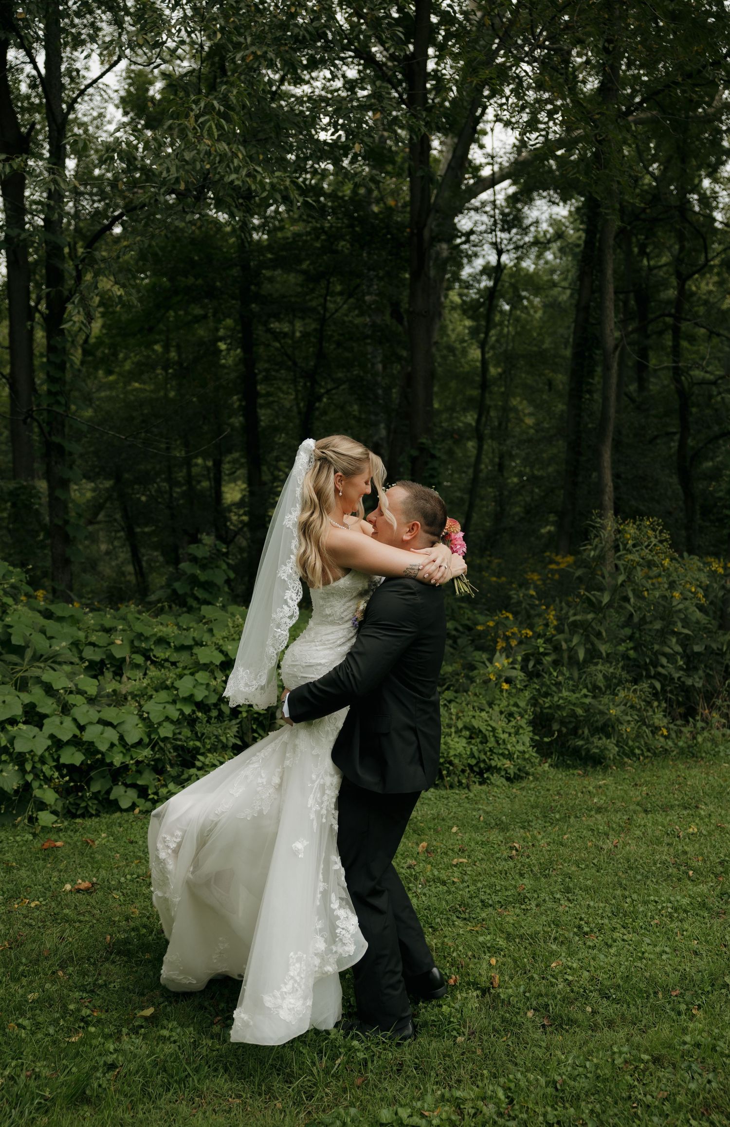 Bride and groom share a romantic kiss as groom lifts bride in a lush green outdoor forest wedding setting.