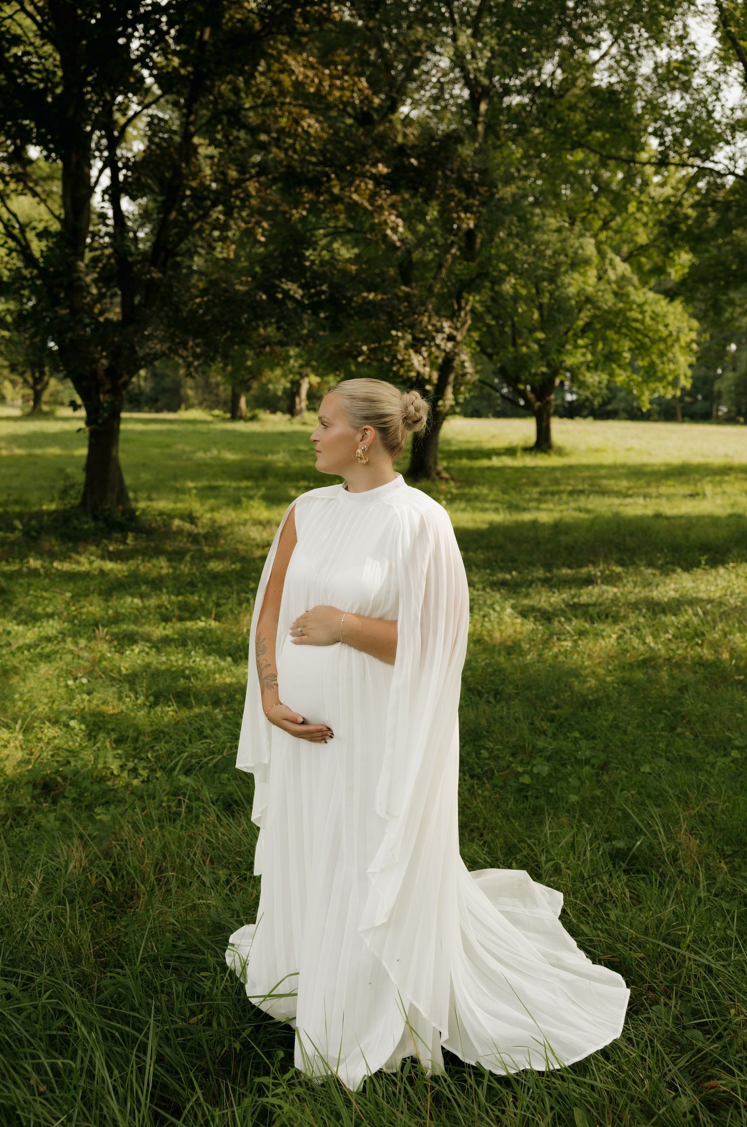 Pregnant woman in flowing white cape gown standing in lush green park, cradling baby bump during outdoor maternity photoshoot.