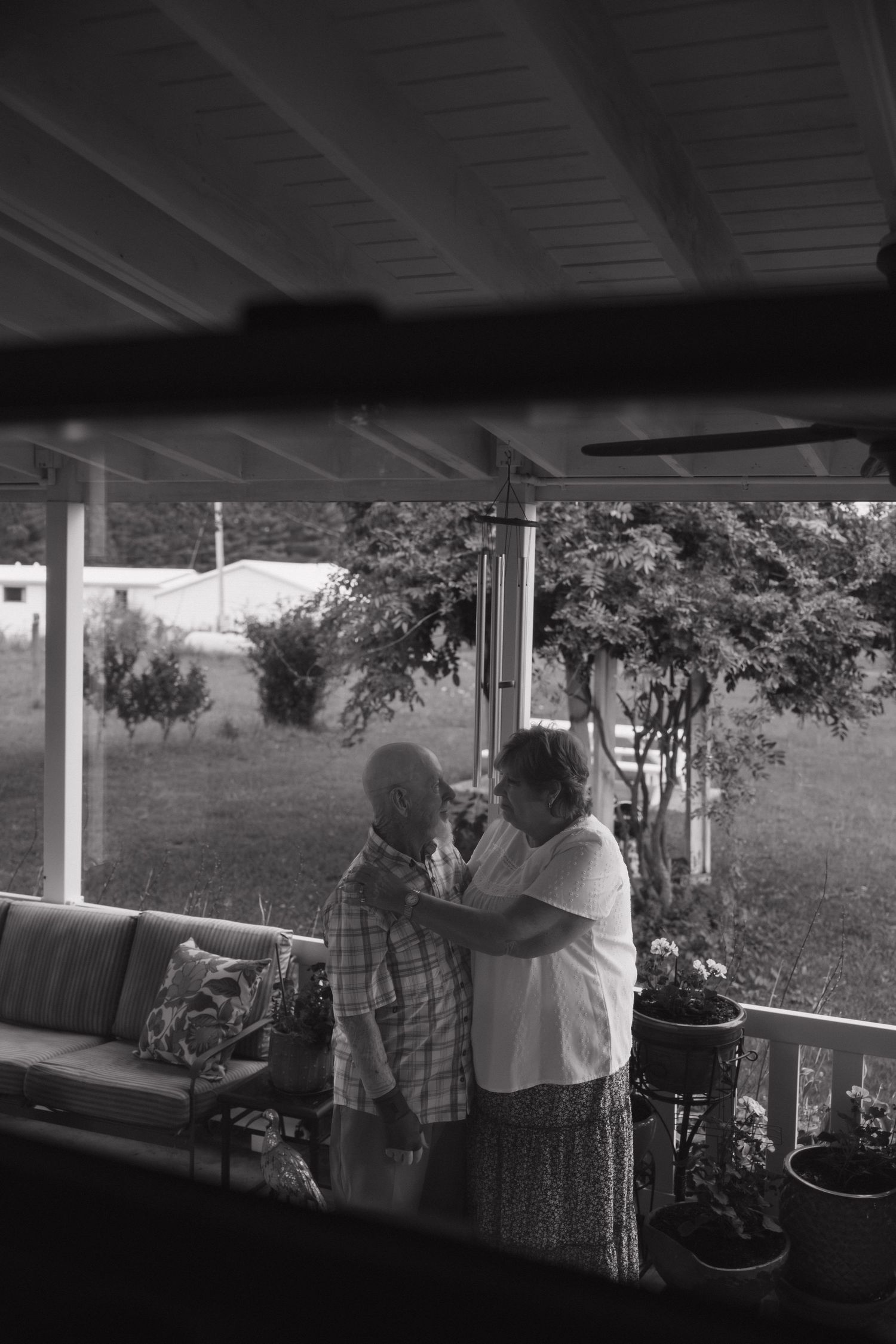 Black and white photo of elderly couple sharing a tender moment on a covered porch surrounded by lush greenery.