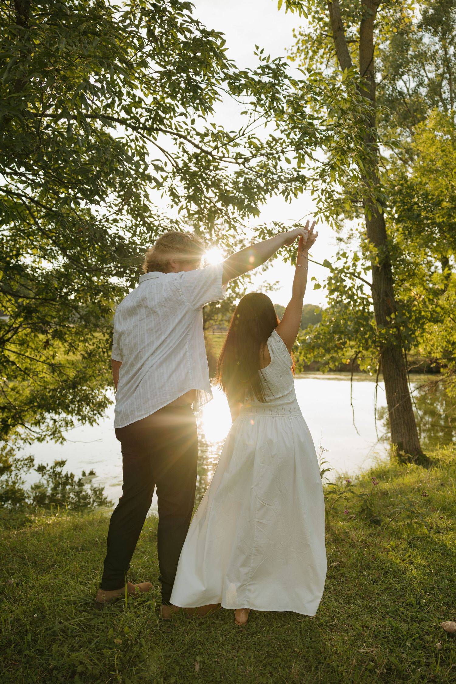 Couple dancing outdoors near a sunlit lake, woman in white dress, man in white shirt, surrounded by lush green trees.