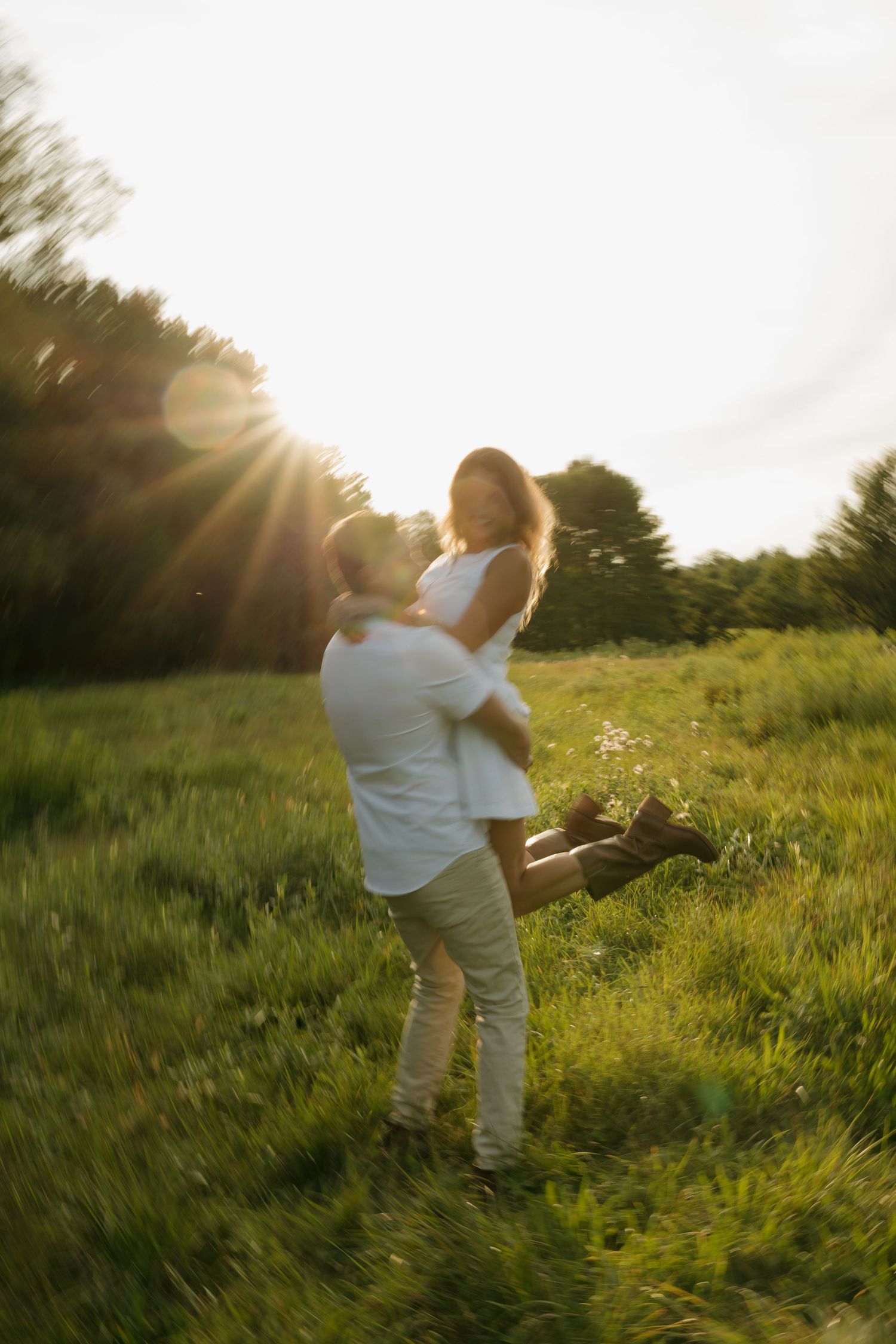 A couple shares a romantic moment in a sunlit green field, as a man lifts a woman in a white dress at golden hour.