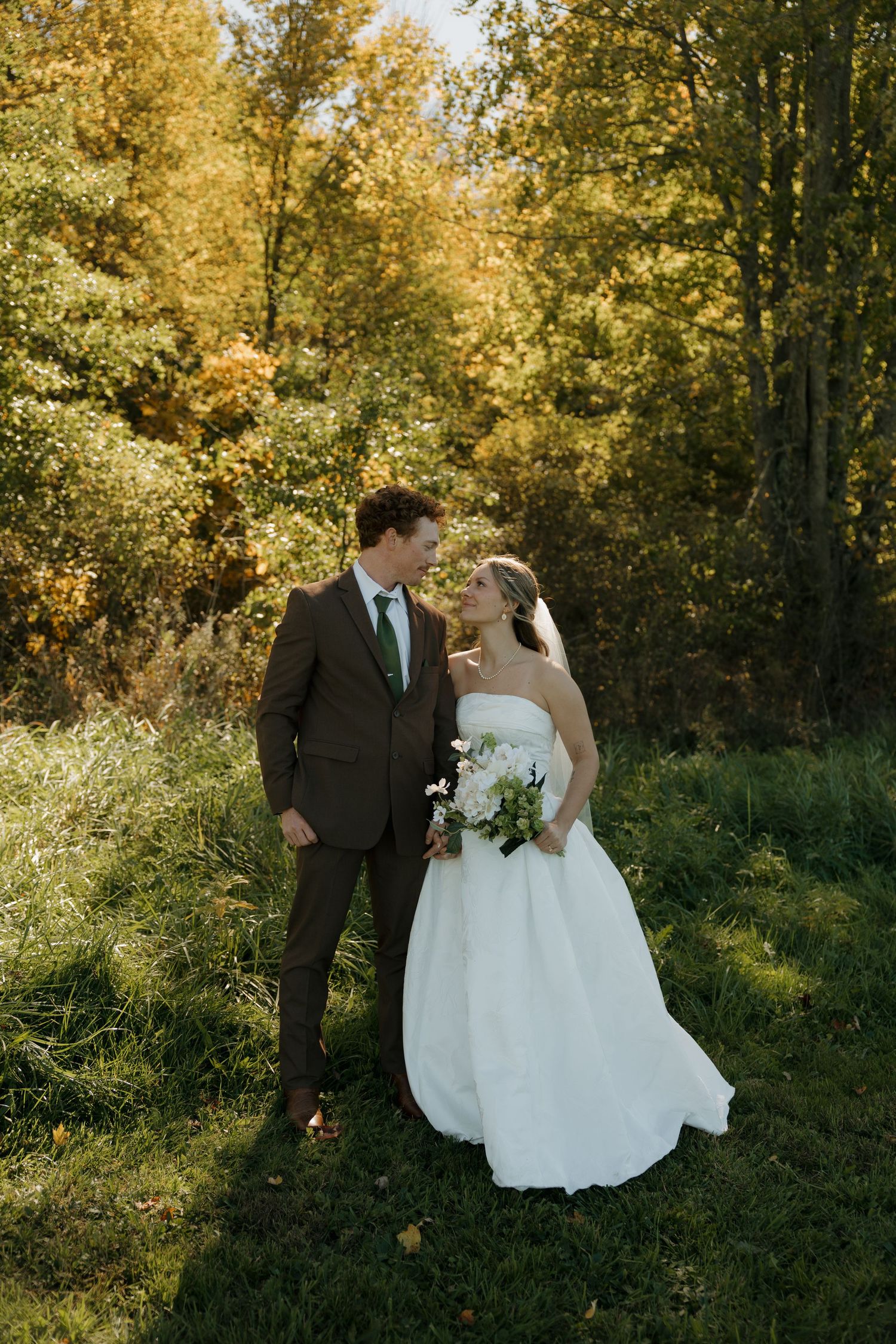 Bride in a strapless white gown holding a bouquet smiles at a groom in a brown suit and green tie in a sunlit autumn forest.
