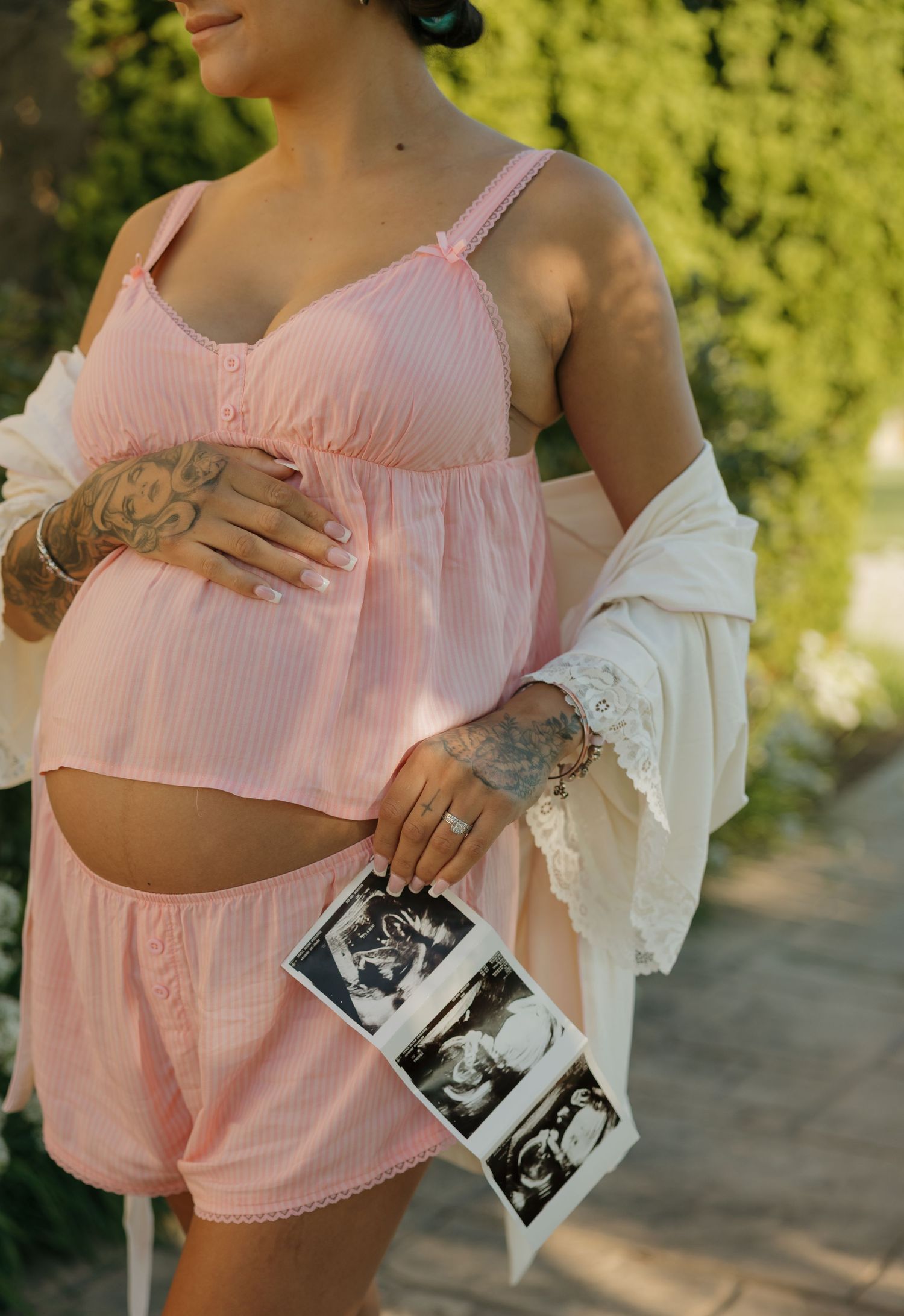 Pregnant woman in pink crop top and shorts holding ultrasound photos, showing baby bump and tattooed arms outdoors.