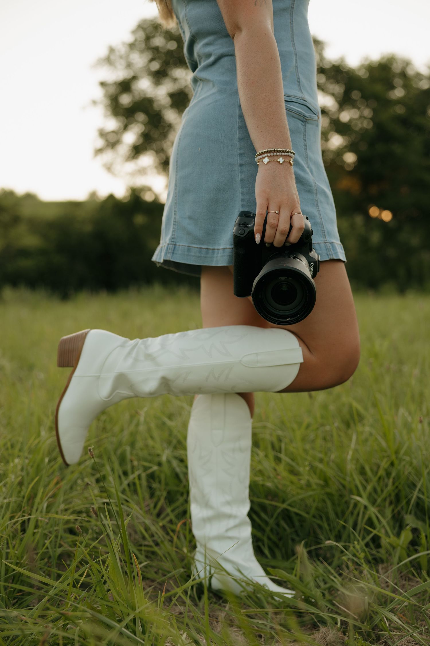 Woman in denim dress and white cowboy boots holding a camera in a green field at sunset.