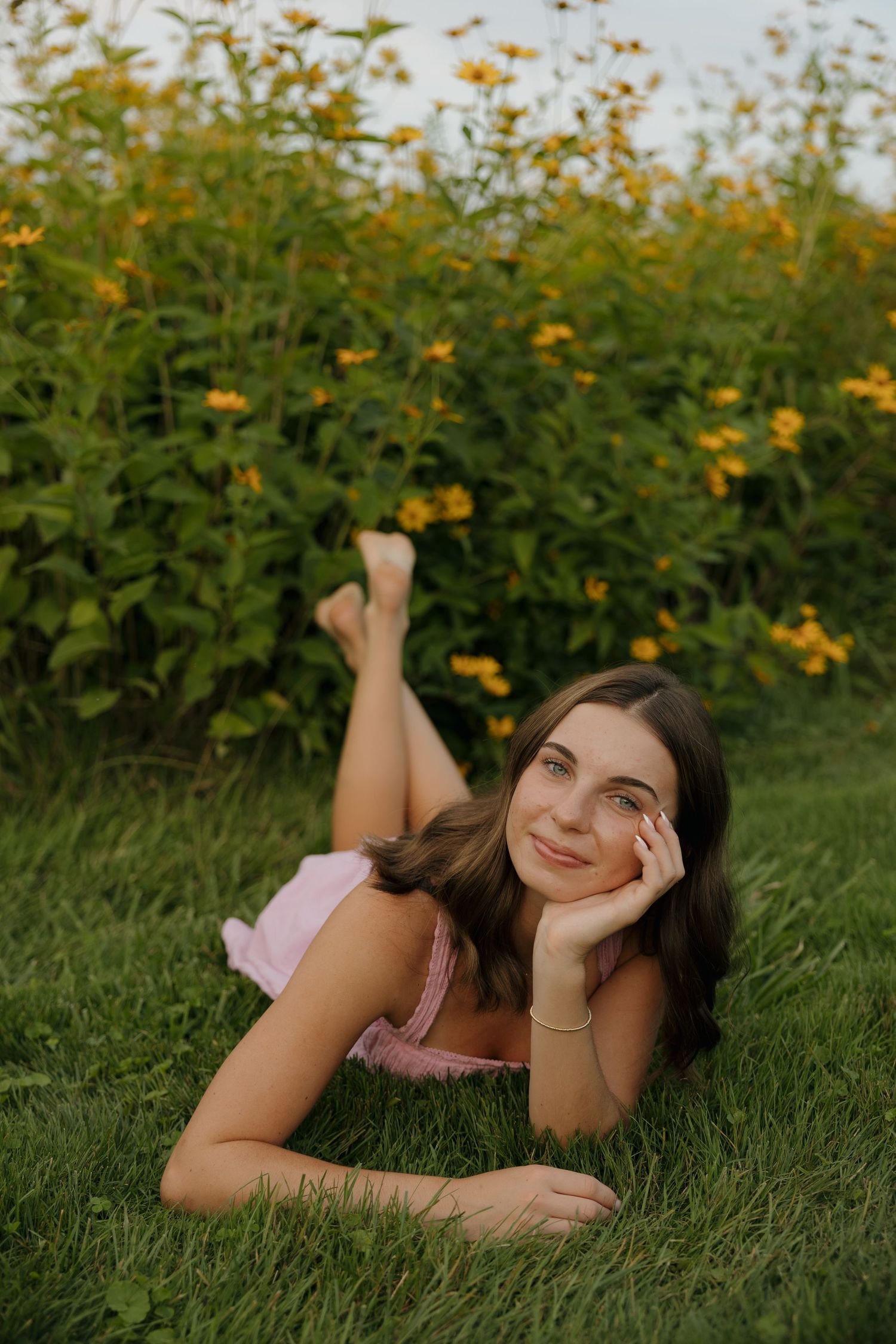 Young woman in pink dress lying on grass smiling, with yellow wildflowers blooming in background.