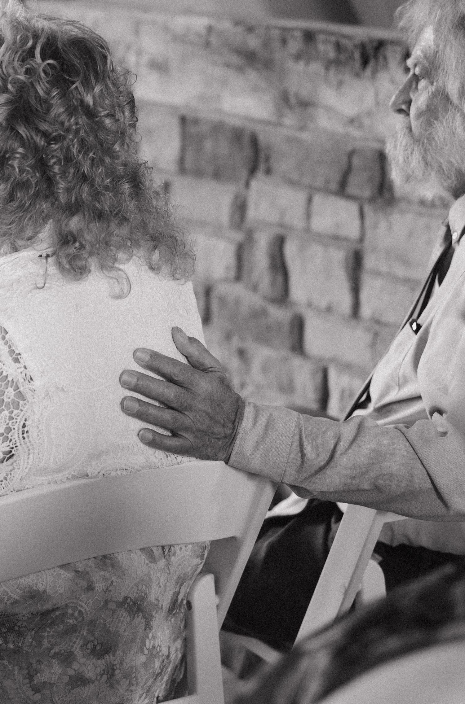 Black and white photo of an elderly bearded man placing his hand on a curly-haired woman's back at a gathering.