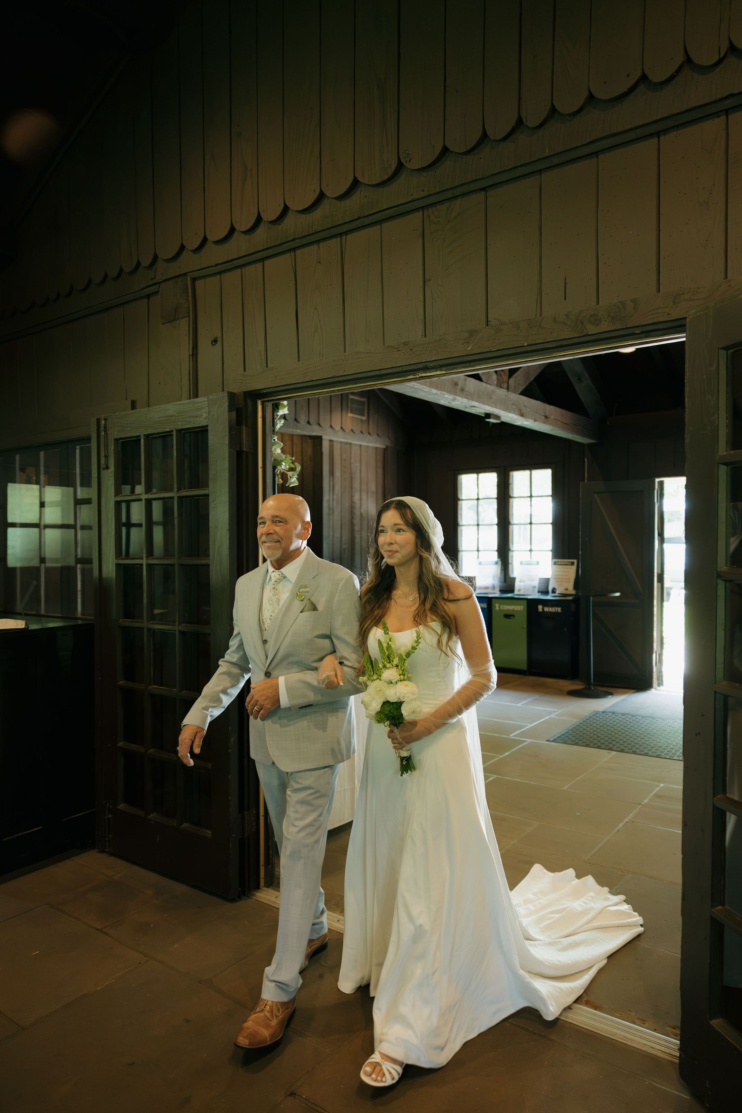 Bride in white gown holding bouquet walks with suited man through rustic barn venue doors on wedding day.