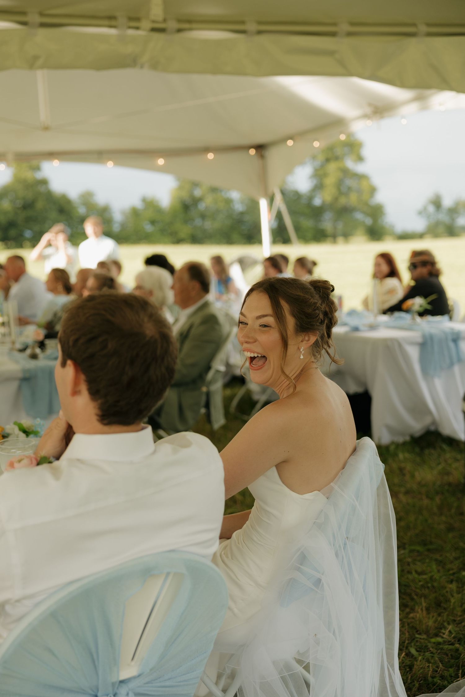 Bride in white dress laughing joyfully at outdoor wedding reception under tent with guests seated at tables.